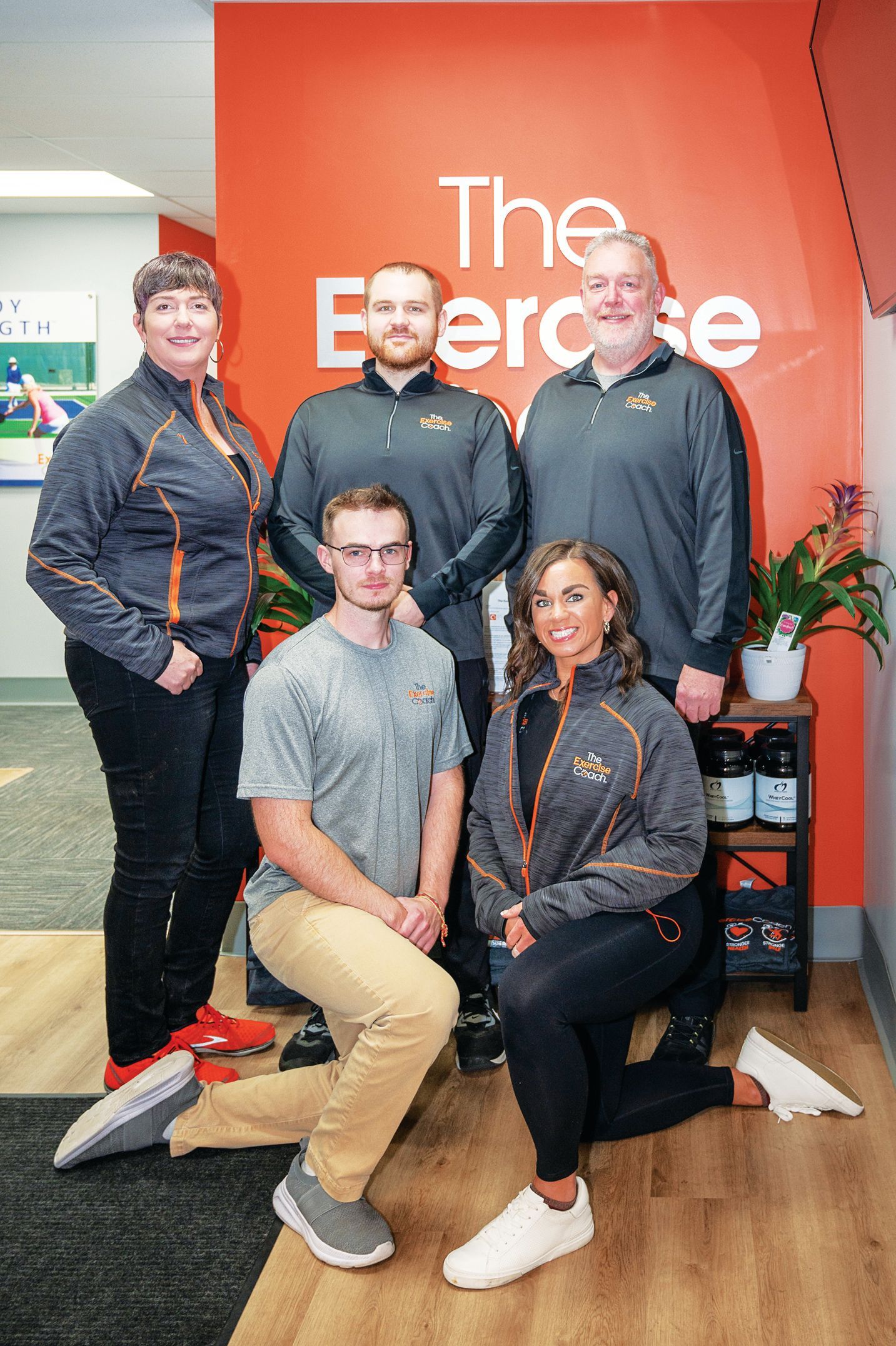 Five people in fitness attire pose in front of an orange wall with 