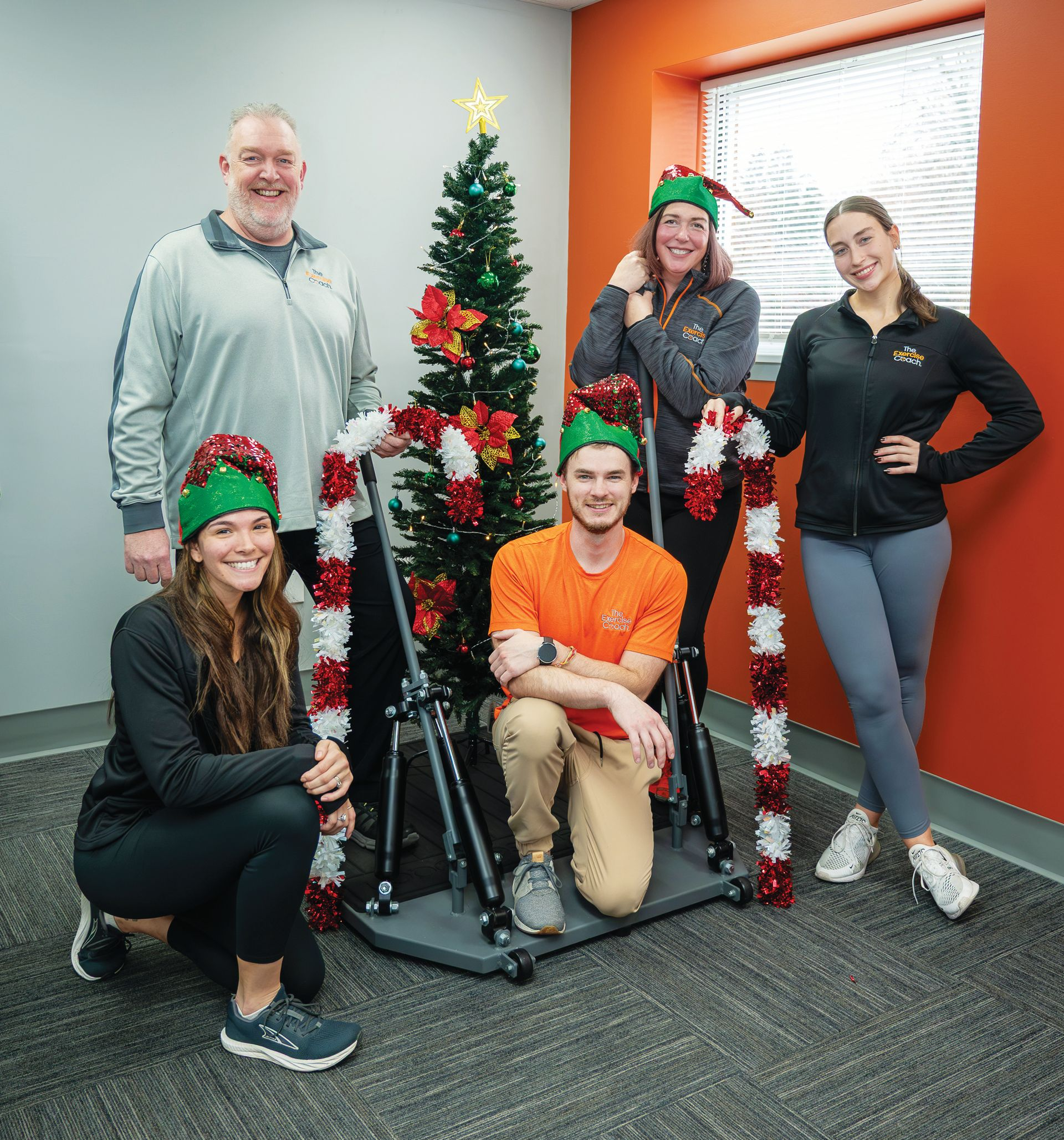 Five people wearing elf hats pose by a decorated Christmas tree in an office with gray and orange walls.
