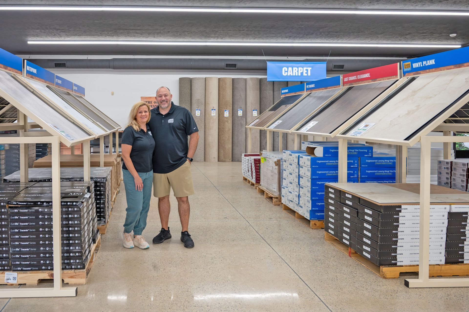 A pair of people posing in a flooring showroom with rows of carpet samples and stacked tile boxes on display.