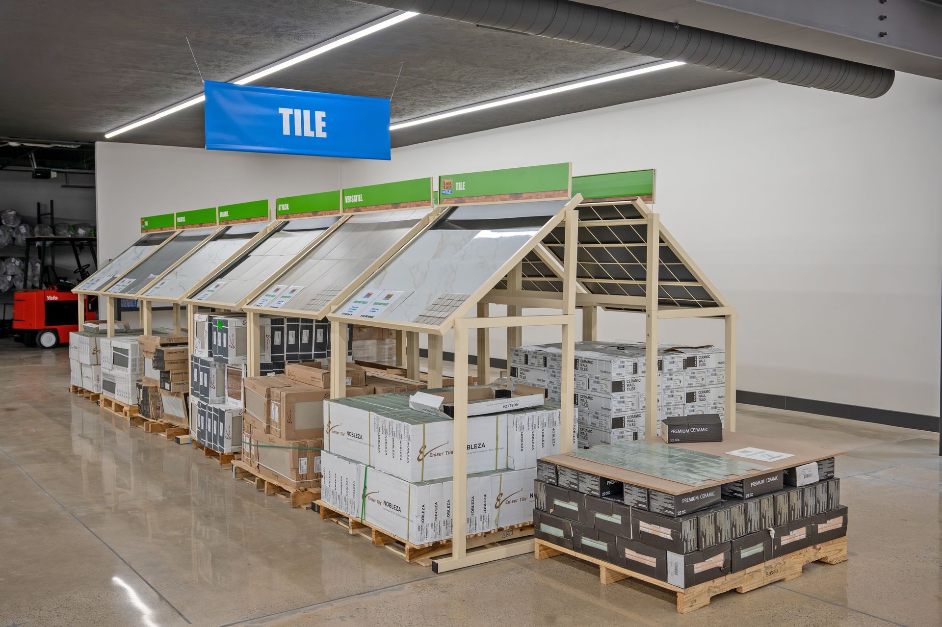 A warehouse tile display rack with sloped panels showing various tile samples above stacked boxes on wooden pallets.