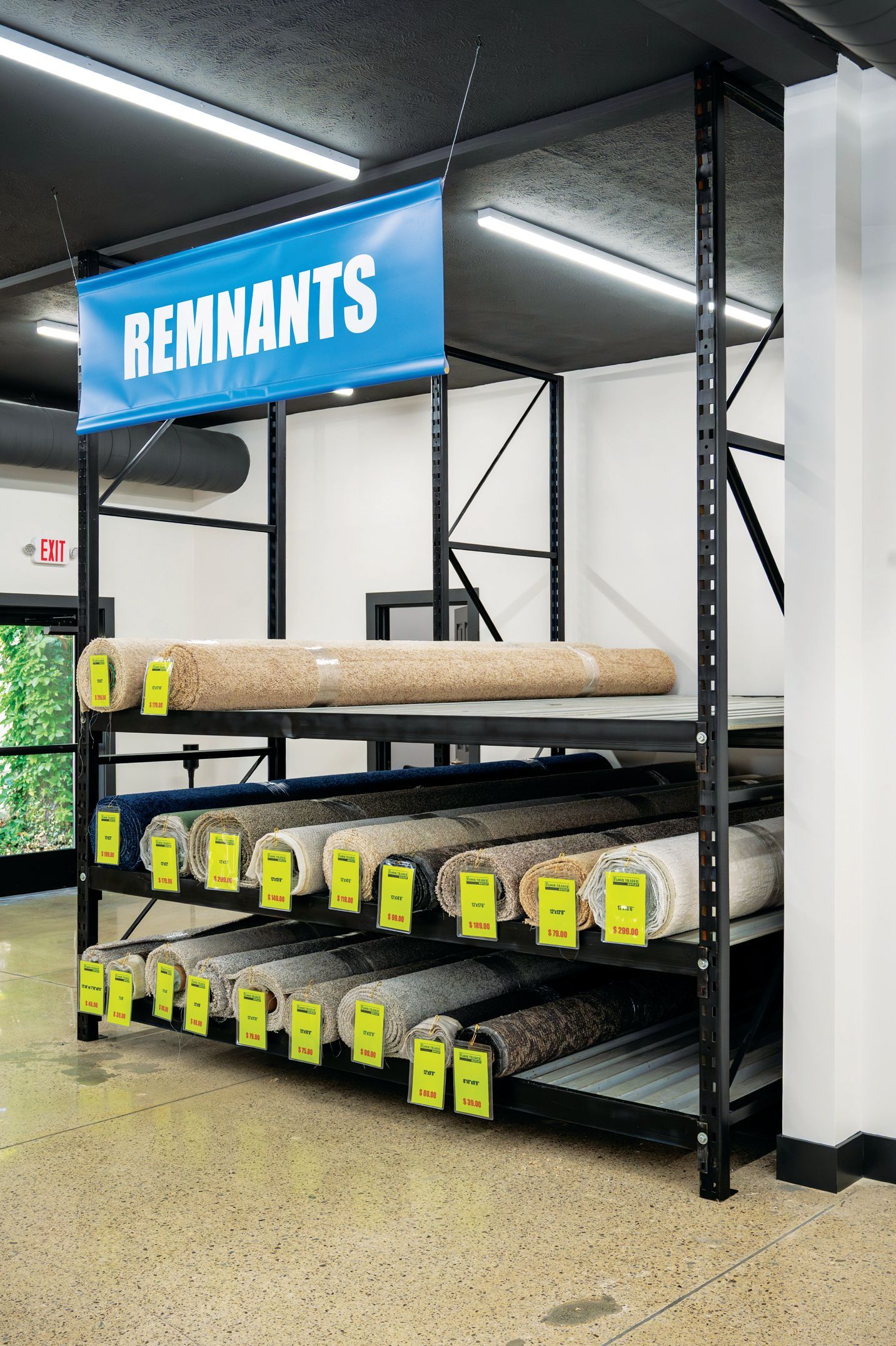 A metal shelving unit in a warehouse holding rolls of carpet, each labeled with a tag, under a sign reading 