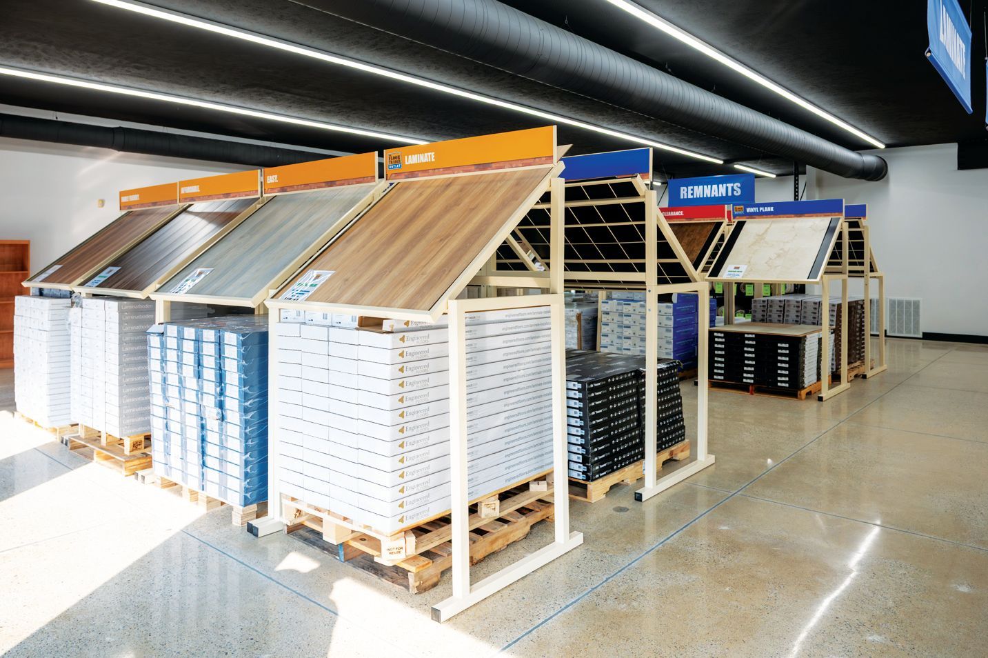 A retail showroom aisle featuring several wooden display racks holding various flooring samples over stacks of boxed tiles.