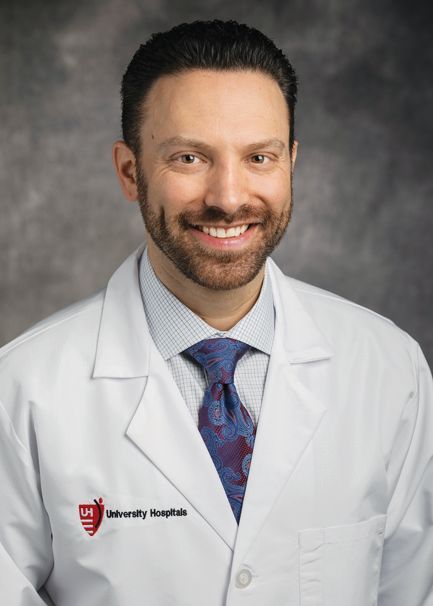 A professional headshot of a smiling medical doctor wearing a white coat with a University Hospitals logo and a necktie.