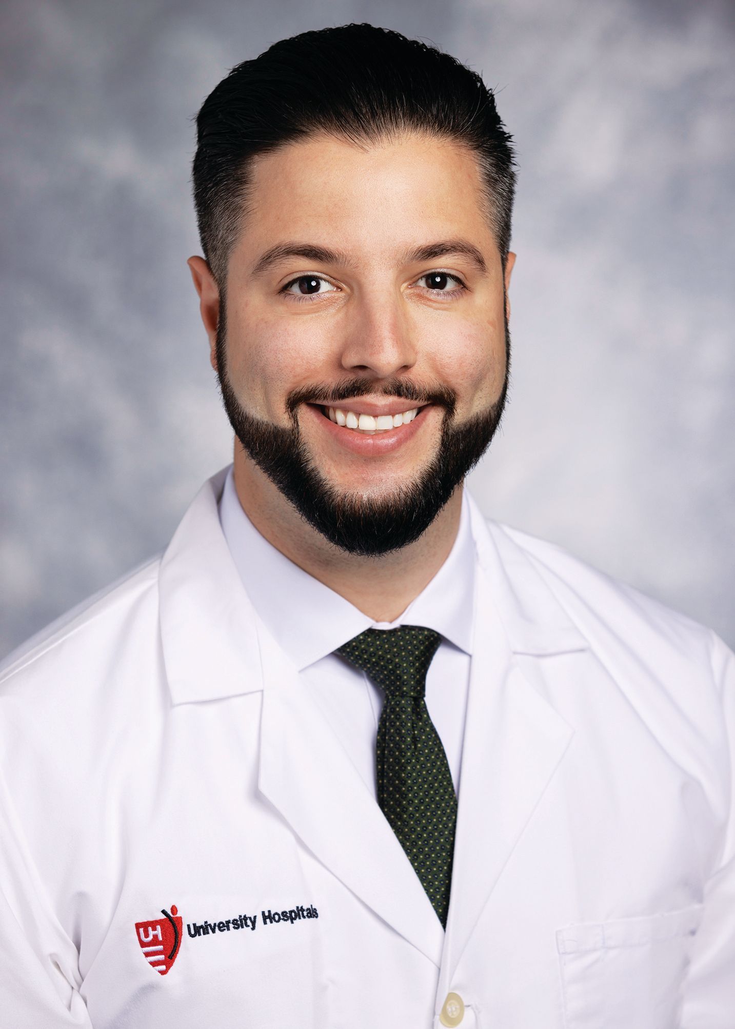 A professional headshot of a smiling medical doctor wearing a white lab coat and a green tie against a mottled background.