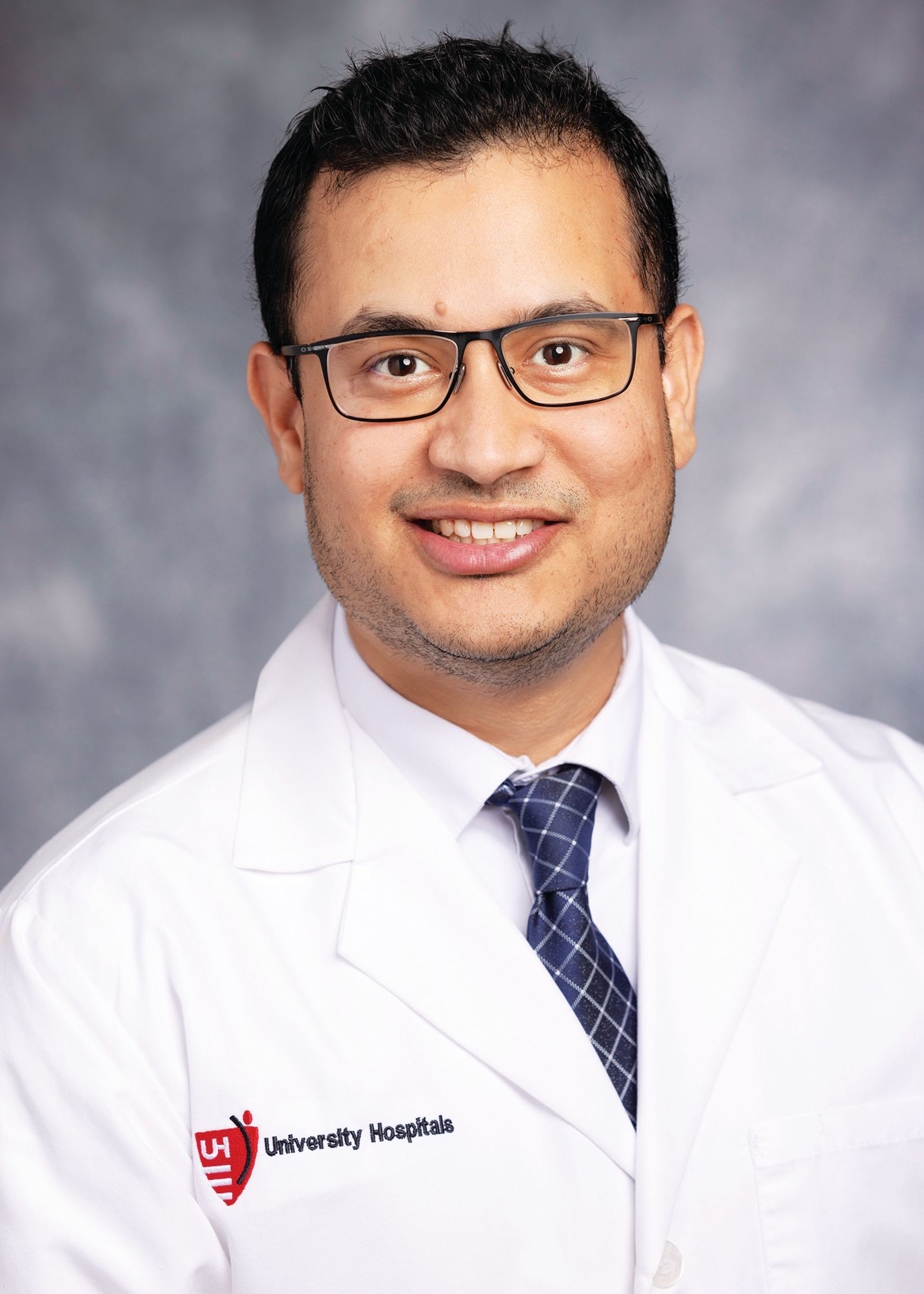 A professional headshot of a person wearing a white medical coat, glasses, and a blue patterned tie against a gray background.