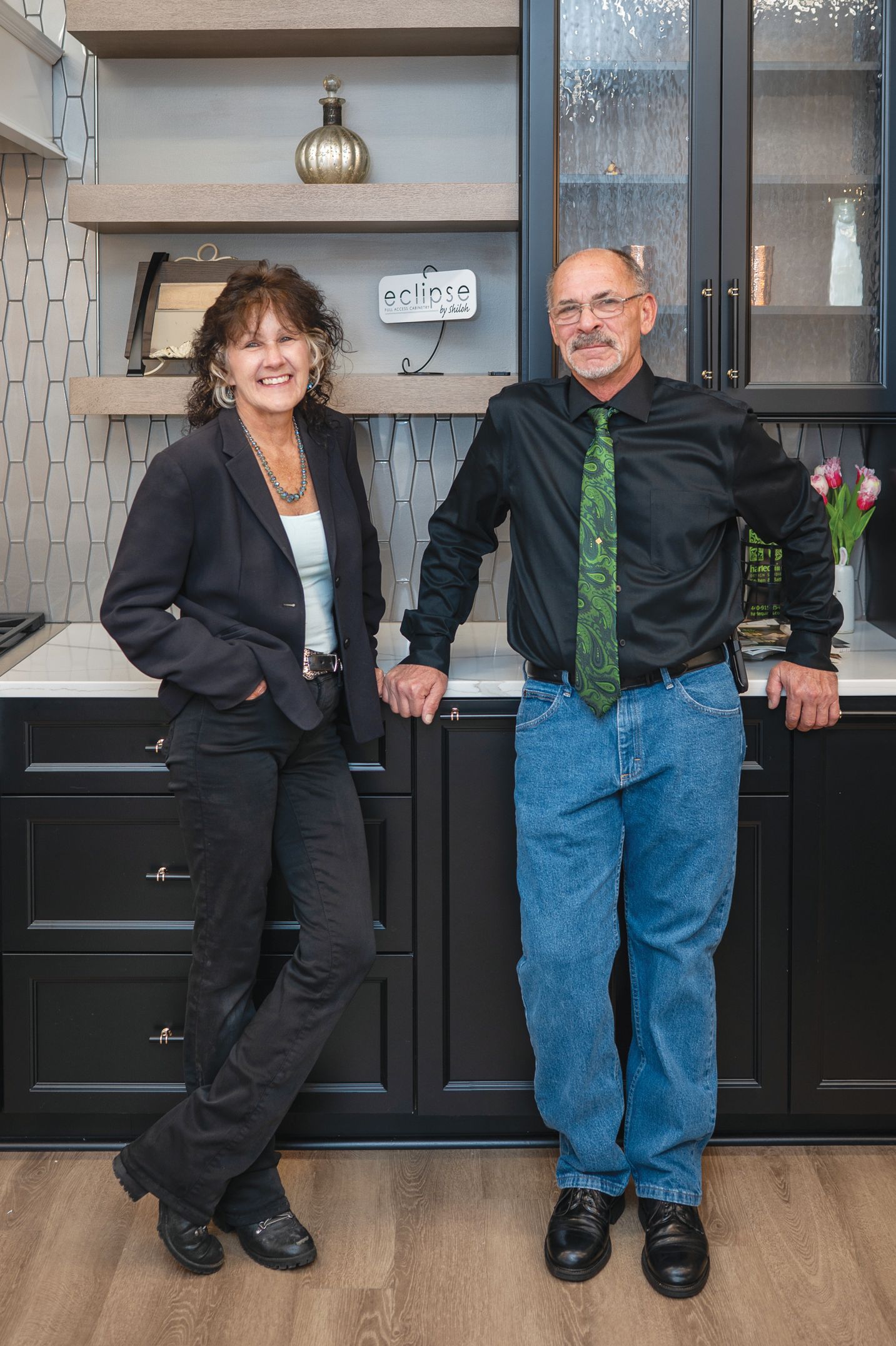 A smiling man and woman stand in a modern kitchen with black cabinets, dark decor, and light wood shelving.