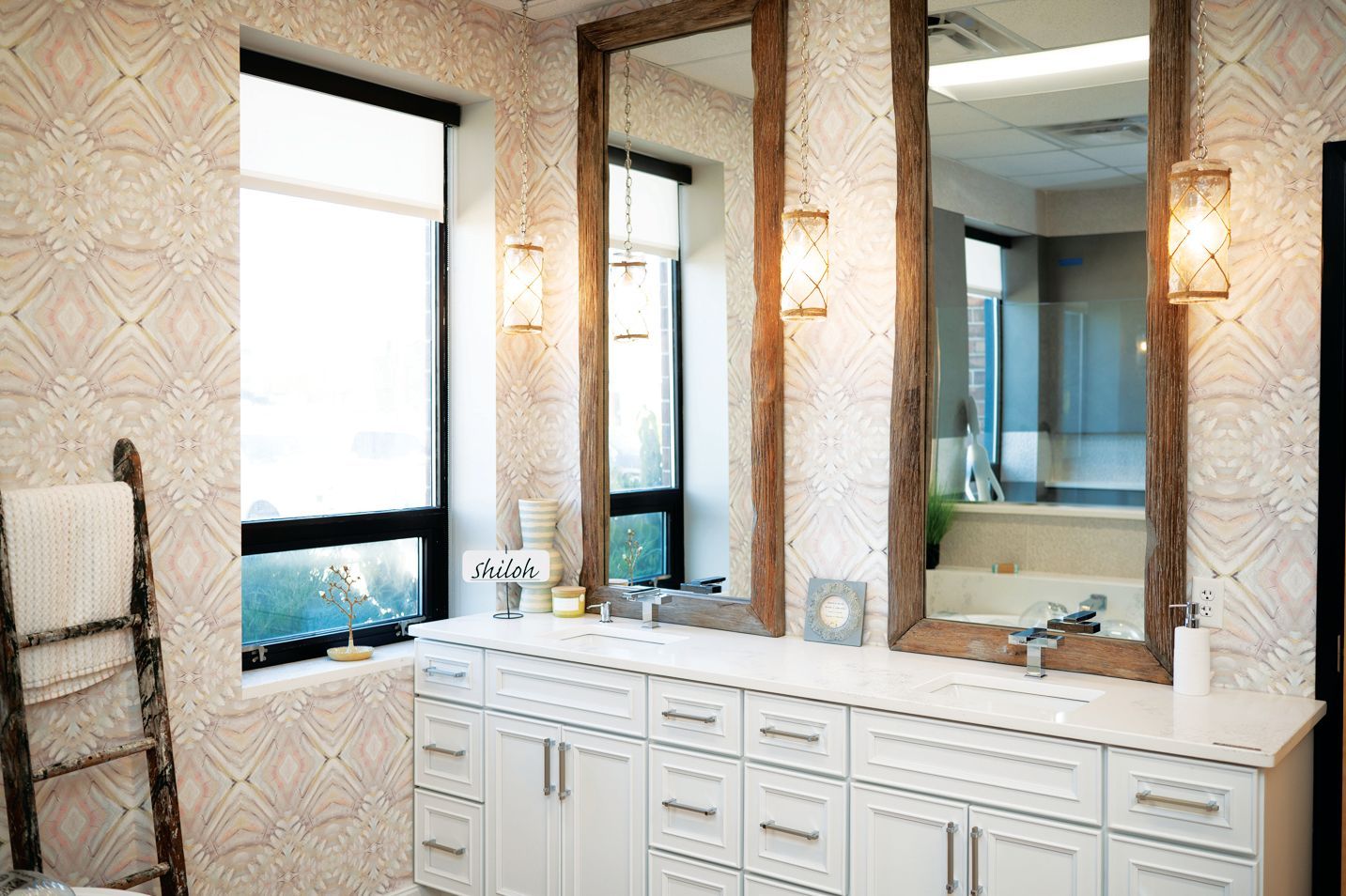 A bright bathroom featuring a white double vanity, large wood-framed mirrors, patterned wallpaper, and a window.