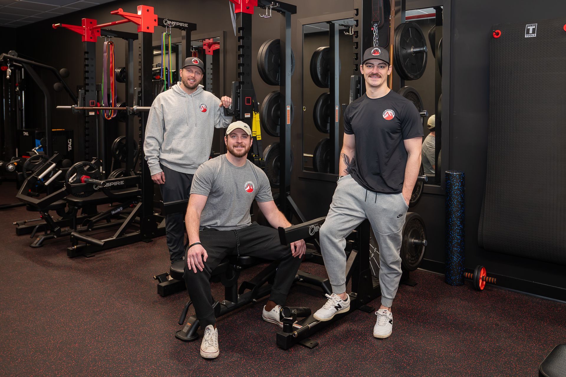 Three people posing in a gym, with one seated on equipment and two standing, all wearing casual athletic apparel.