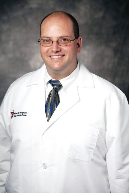 A person in a white lab coat and tie, smiling, posed against a gray backdrop.