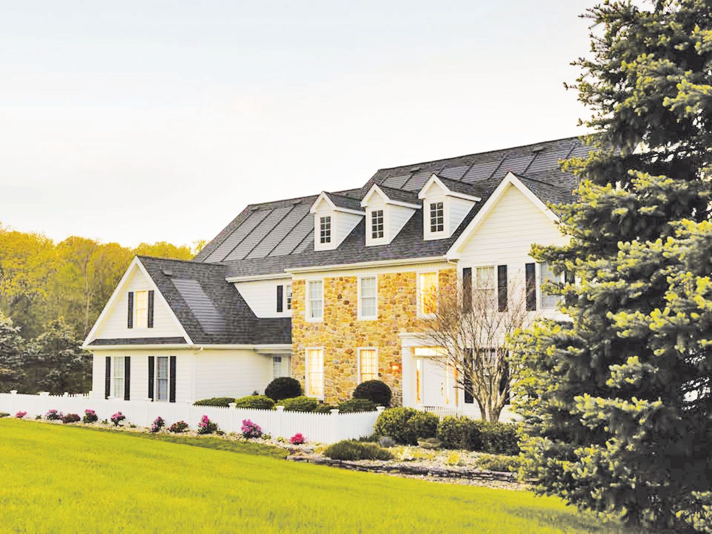 A large, two-story house with white siding and a stone facade, situated on a green lawn with a white picket fence.