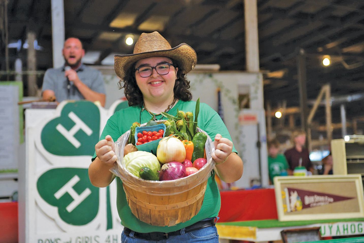 A person in a straw hat and green shirt holds a basket of fresh produce at a 4-H event, with a speaker in the background.