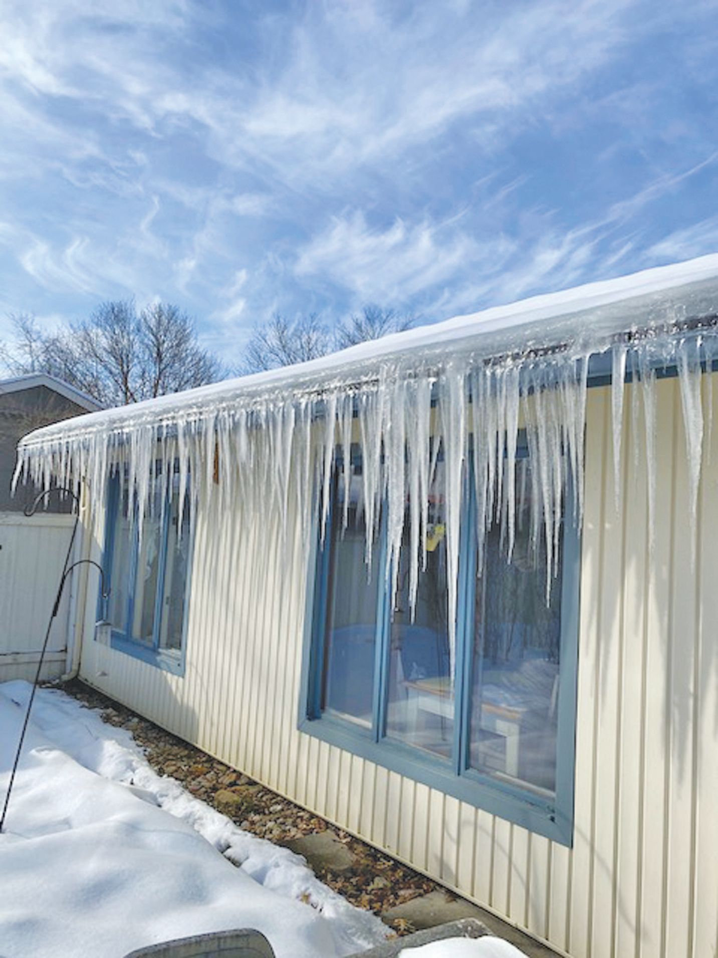 Long, sharp icicles hang from the cream-colored siding of a house under a bright, cloudy blue sky.