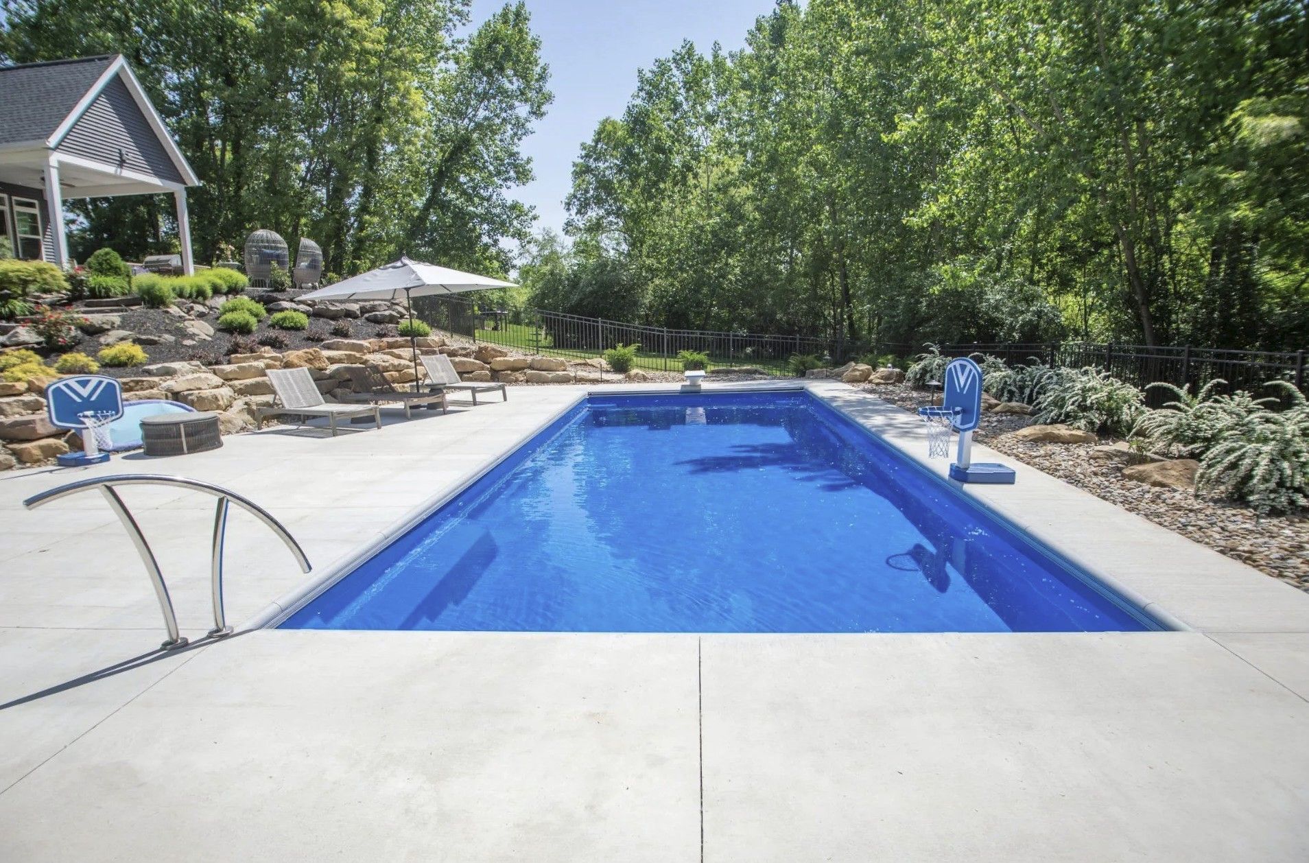 A rectangular, blue swimming pool set in a sunny backyard with concrete decking, lounge chairs, and a retaining wall.