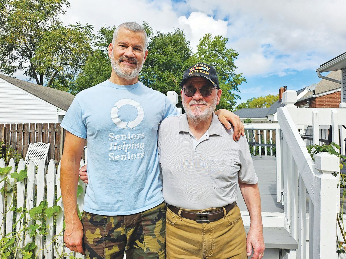 Two men standing side-by-side outside on a sunny day, smiling at the camera with a white fence and deck in the background.