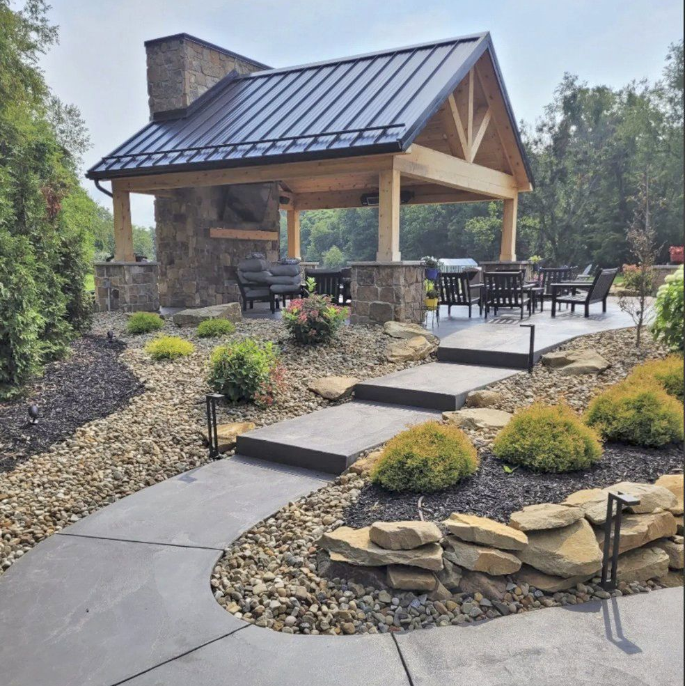 A stone walkway leads to an outdoor timber-frame pavilion with a metal roof, stone fireplace, and patio seating area.