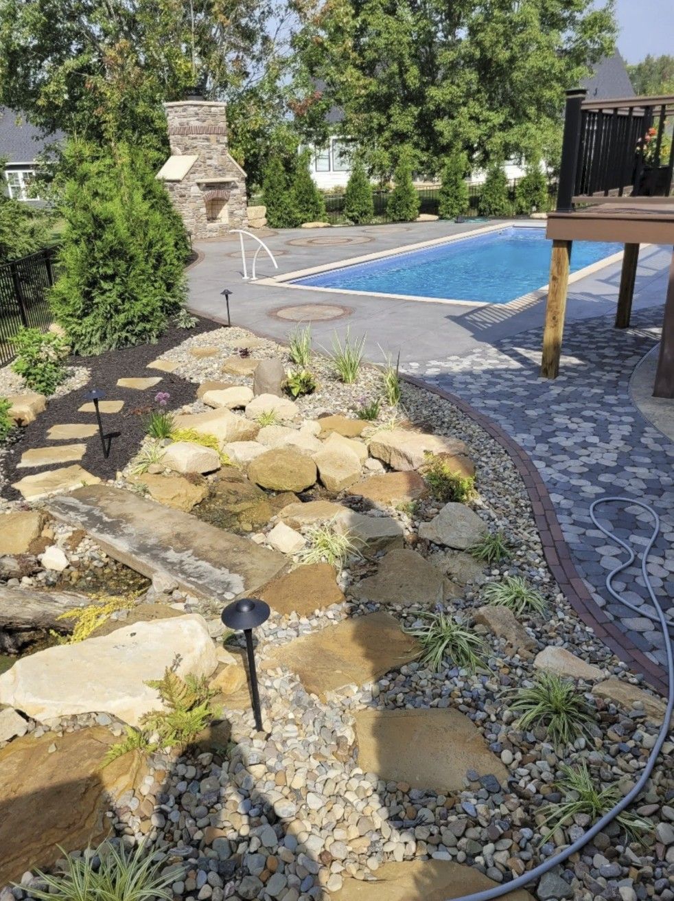 A backyard landscape features a pool, a stone fireplace, and a stone-lined garden area with stepping stones and plants.