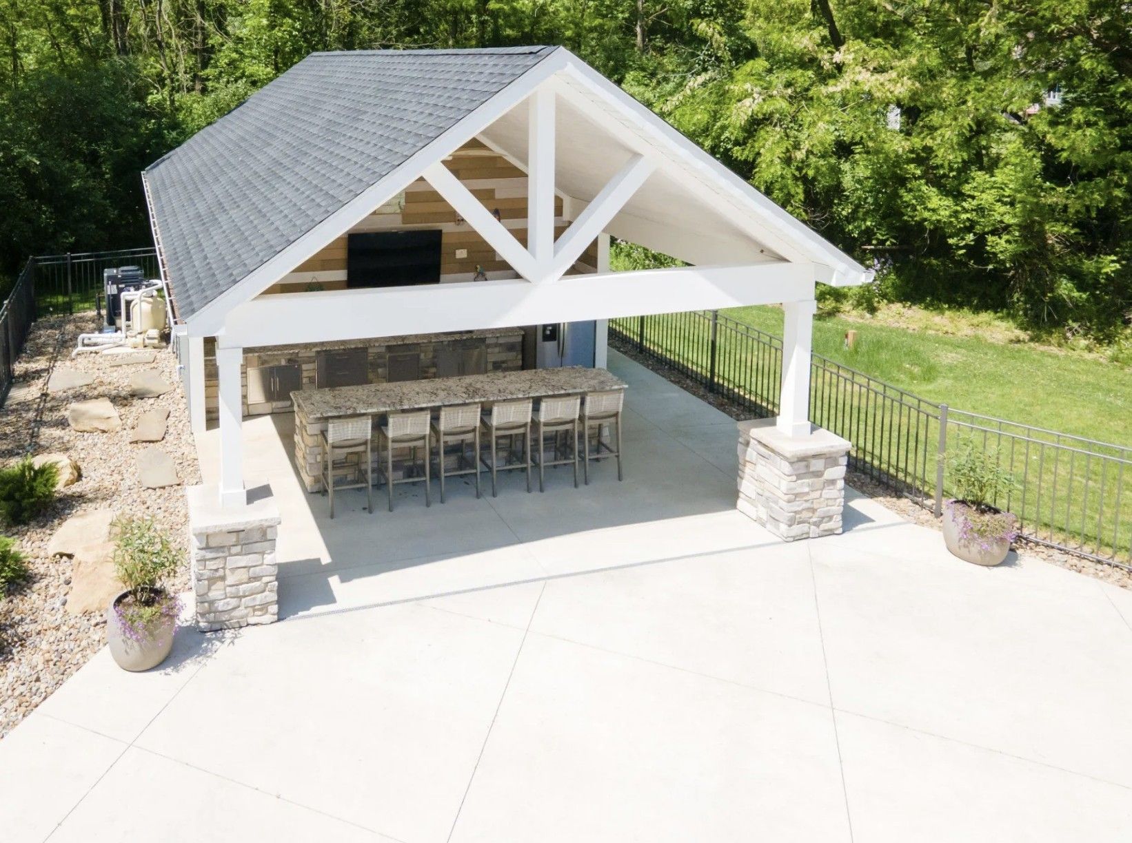 An outdoor covered patio with a stone bar, seating for six, and a mounted television, set in a sunny backyard.