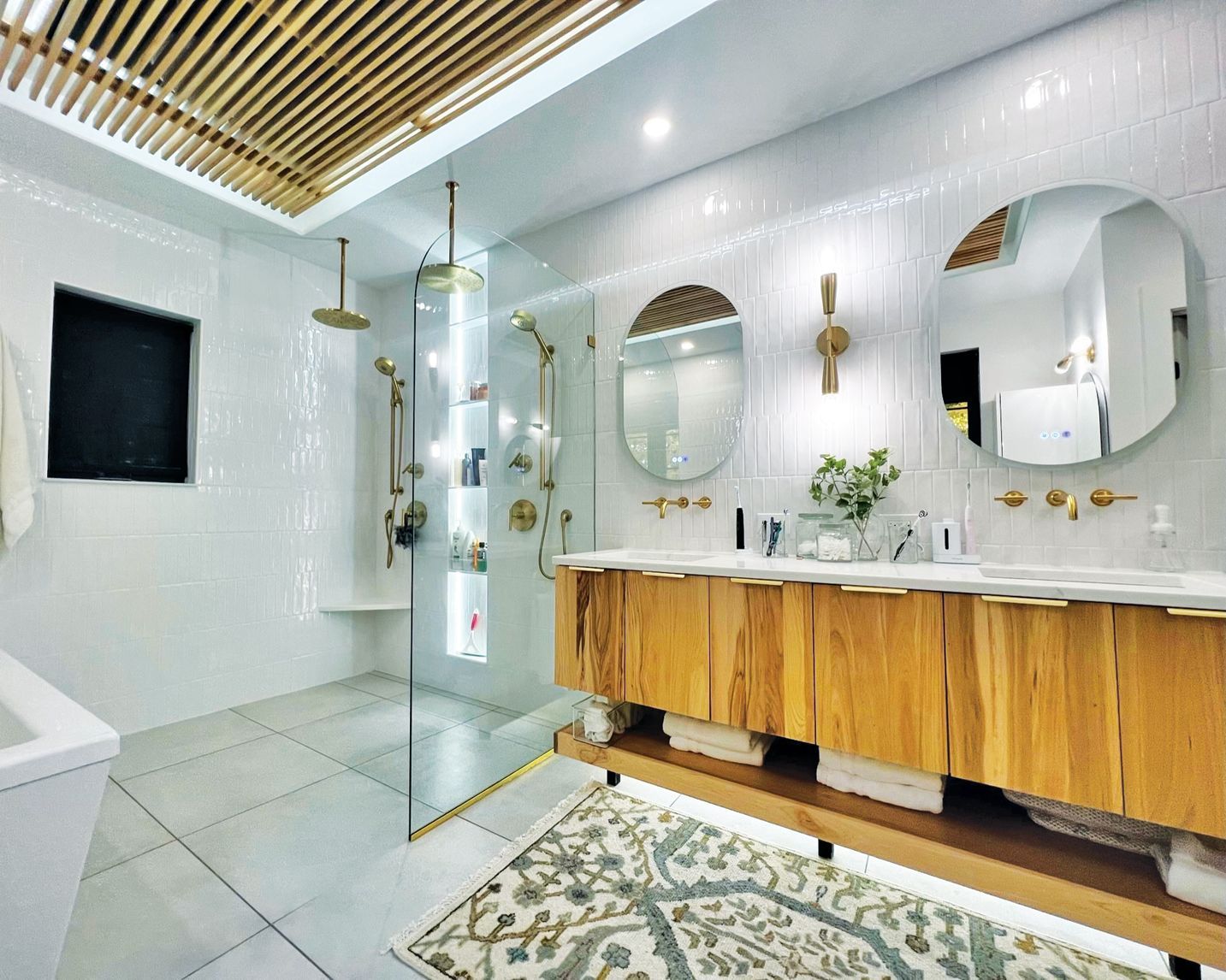 A bright, modern bathroom featuring a wooden double vanity, two round mirrors, a glass walk-in shower, and a slatted ceiling.