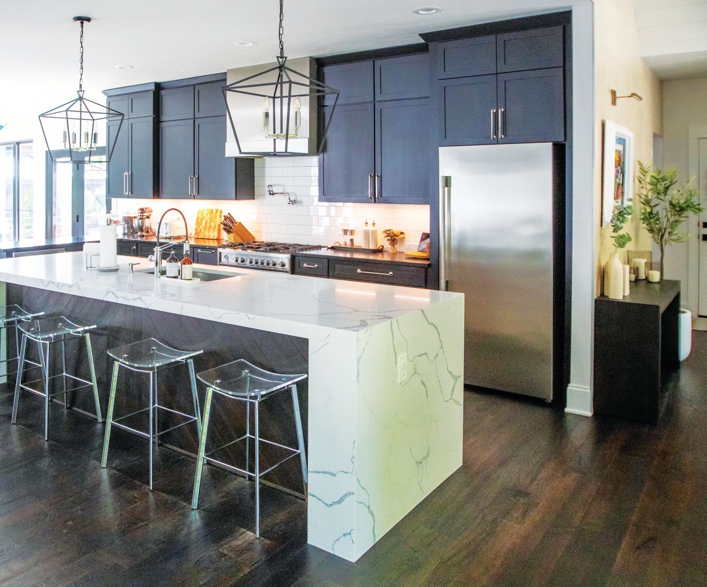 A modern kitchen featuring navy blue cabinets, a marble waterfall island with three stools, and stainless steel appliances.