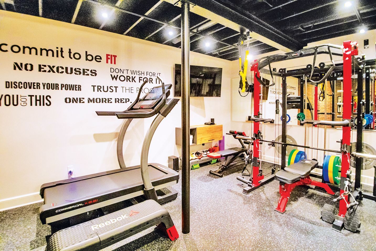 A home gym with a treadmill, red squat rack, weights, and motivational wall text in a room with a black ceiling.