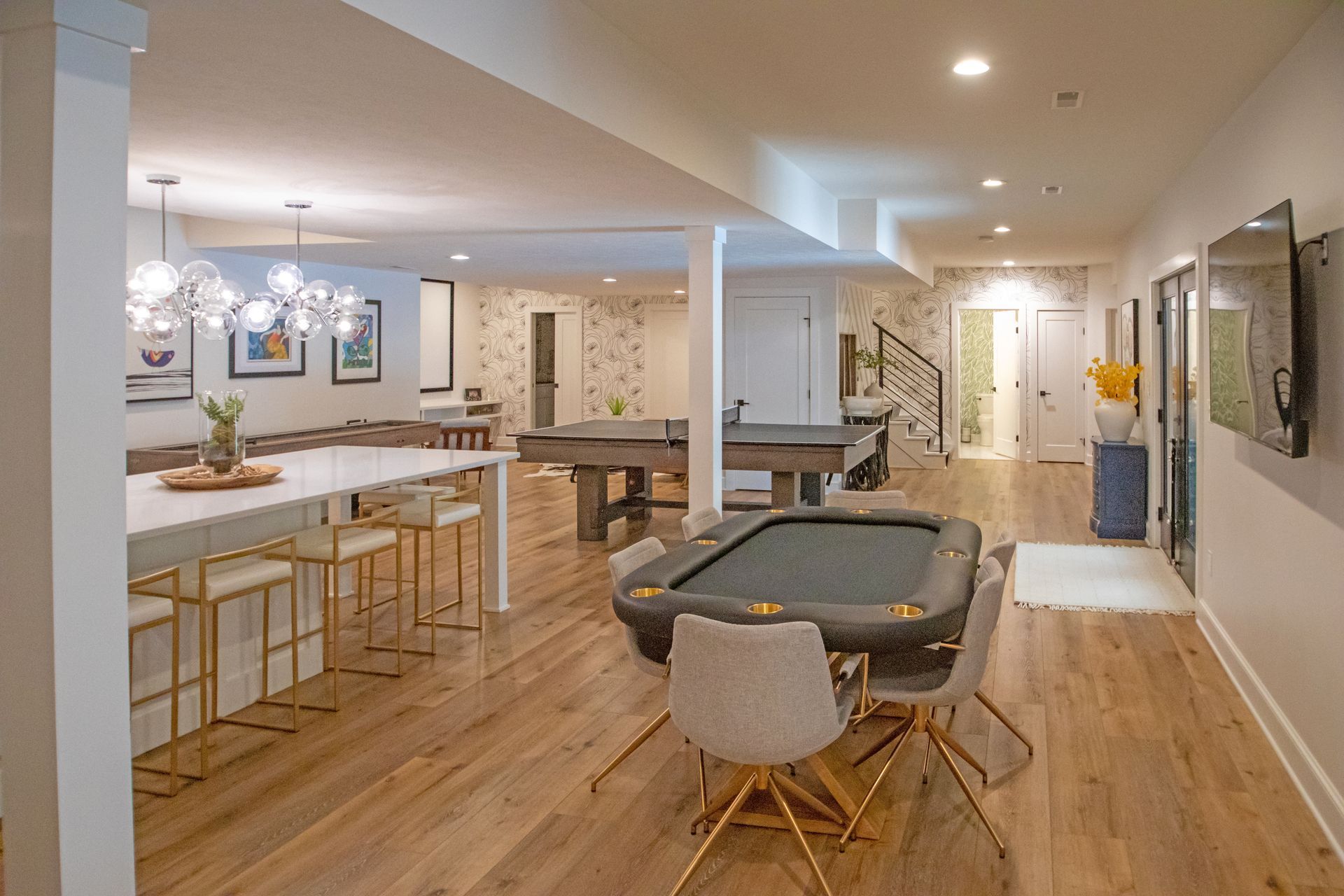 A bright, modern basement with a kitchen island, poker table, and pool table on light wood floors.
