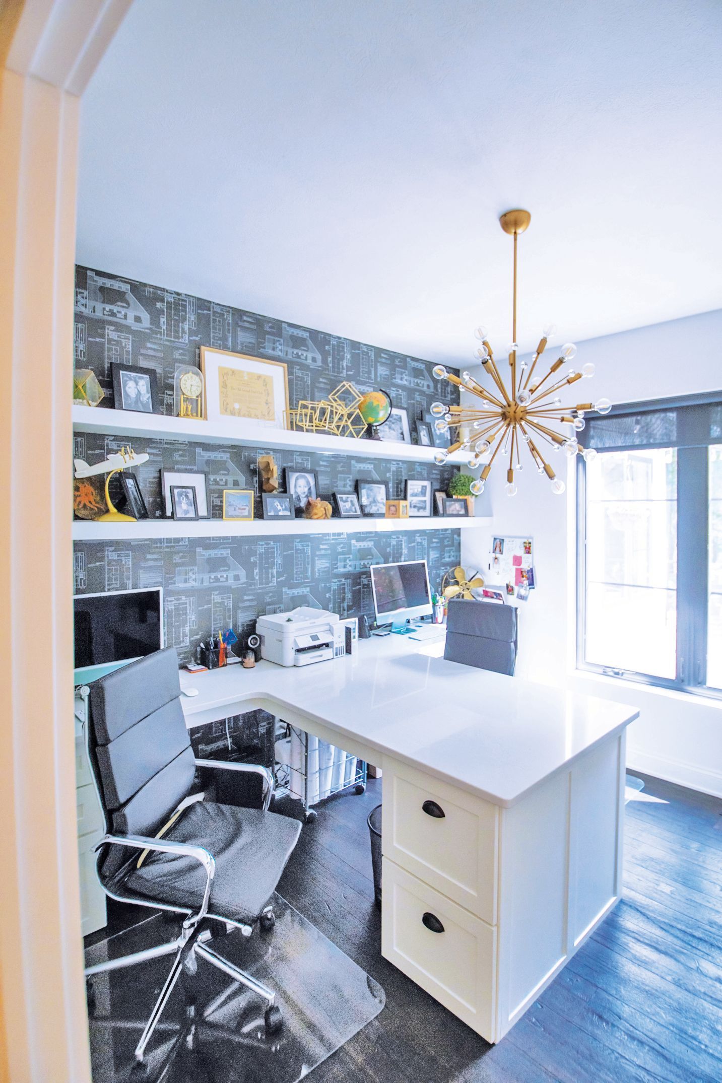 A bright home office features a white L-shaped desk, a dark gray office chair, floating shelves, and a gold sputnik lamp.