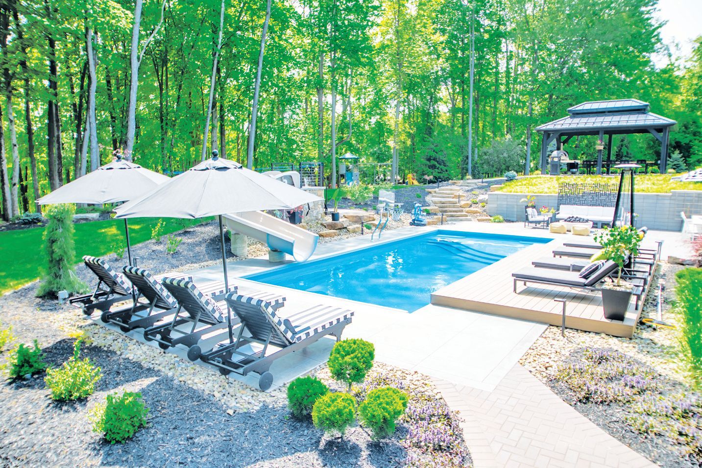 A backyard swimming pool with lounge chairs, umbrellas, a slide, and a wooden patio under a gazebo in a wooded area.