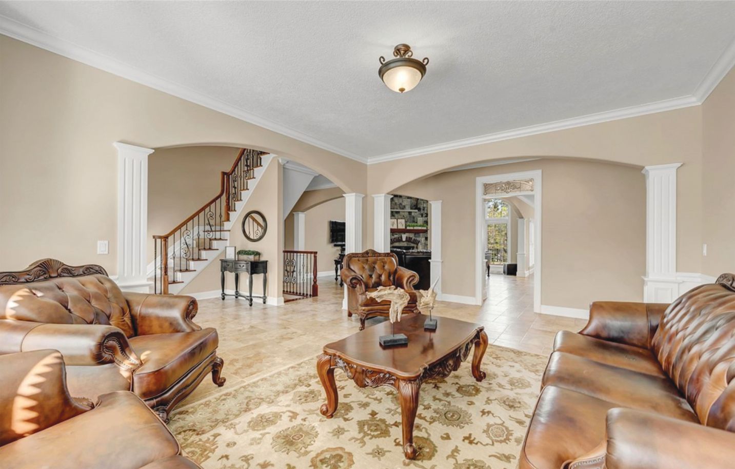 A formal living room with tan walls, brown leather furniture, a patterned rug, and a staircase in the background.