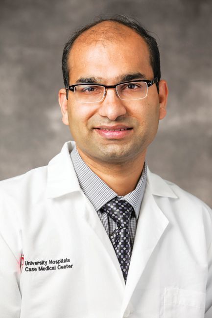 A professional portrait of a doctor wearing a white coat, a patterned tie, and glasses against a grey background.