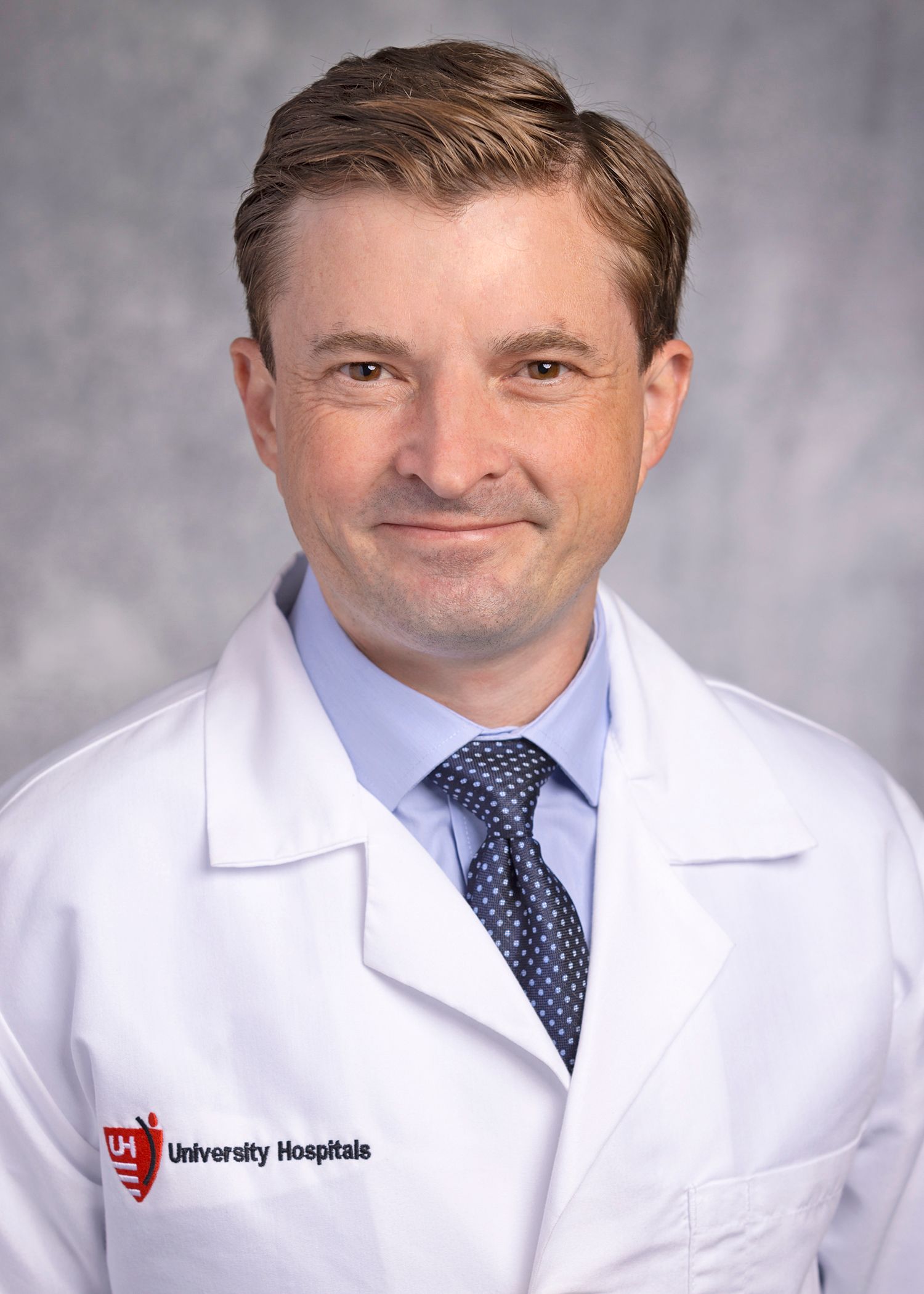 A professional headshot of a person in a white medical coat and a blue dress shirt with a polka-dot tie.