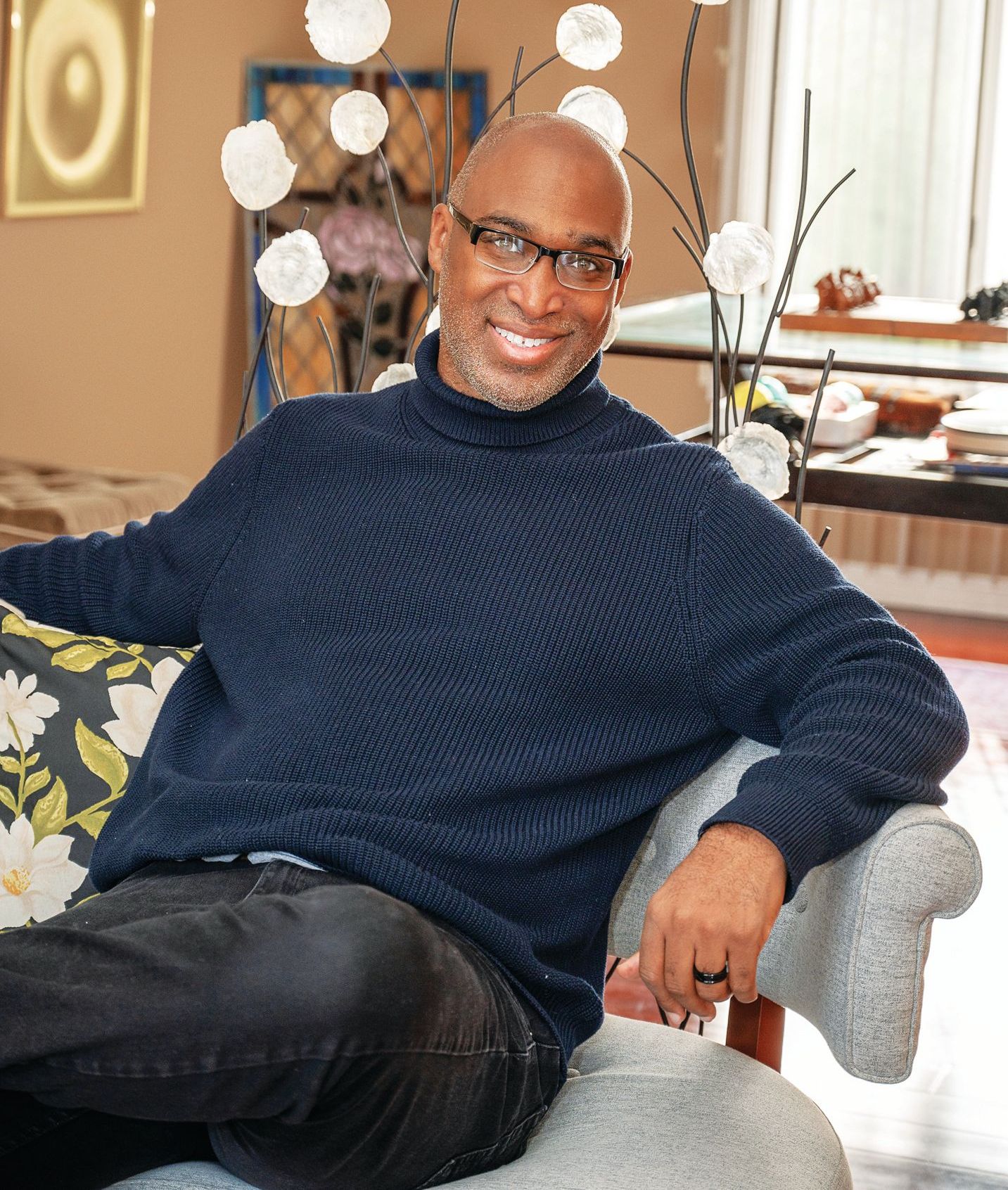 A smiling man wearing a dark blue turtleneck sweater sits on a patterned sofa in a room with decorative branches.