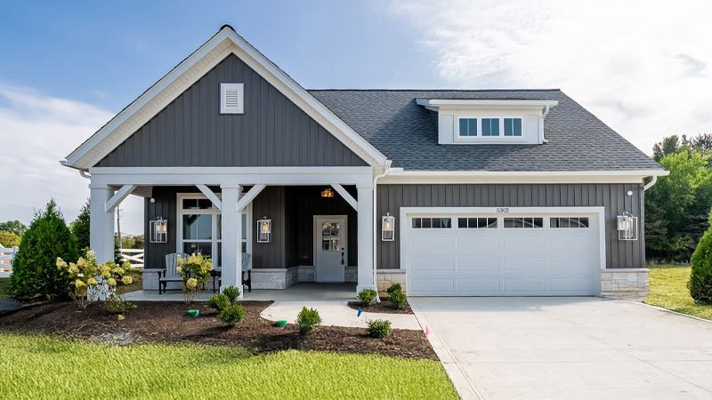 A one-story gray house with white trim, a front porch, and a two-car garage sits on a grassy lot under a blue sky.