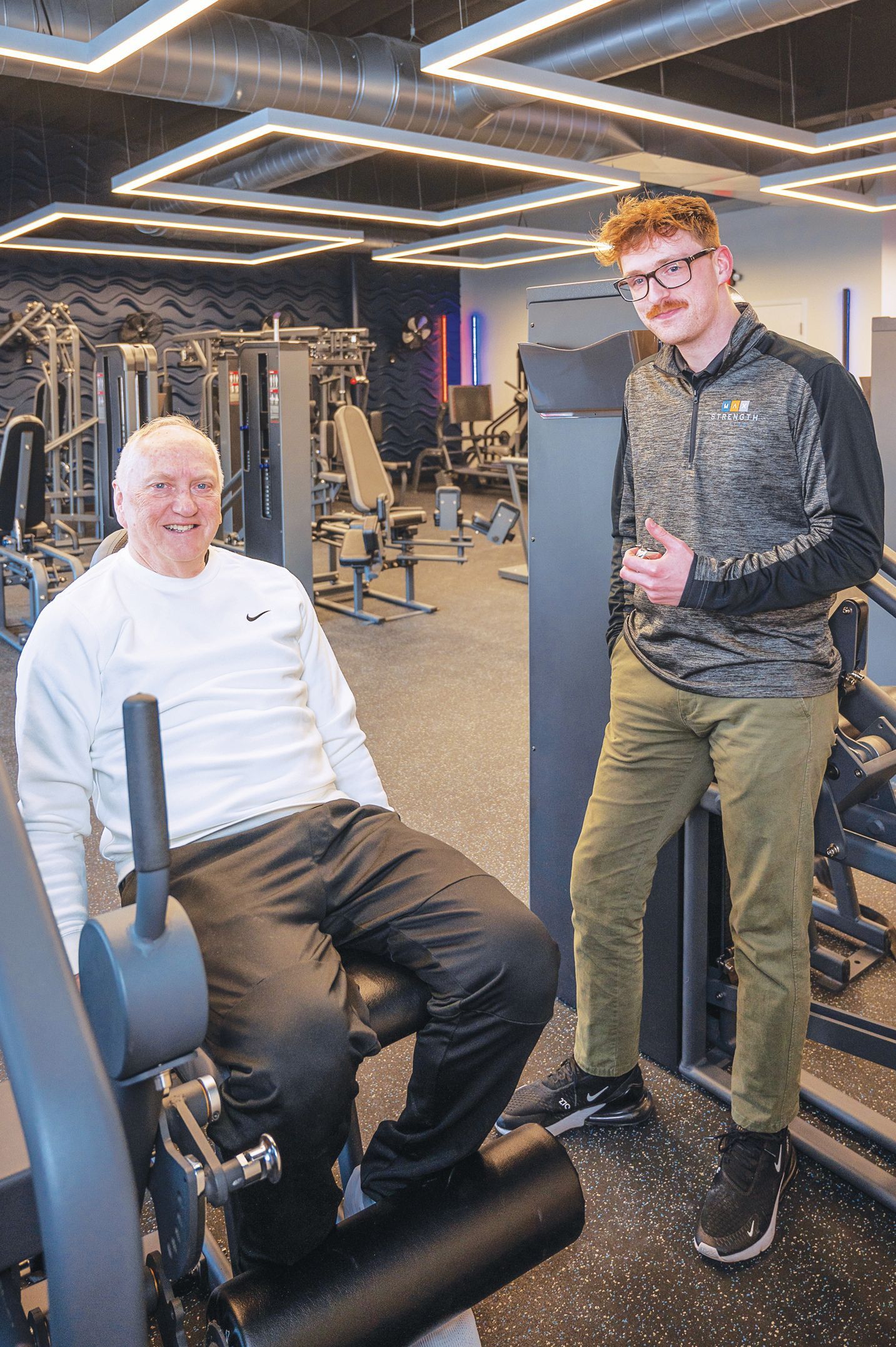 A man sitting on a gym machine smiles next to another person standing by a weight station in a fitness center.