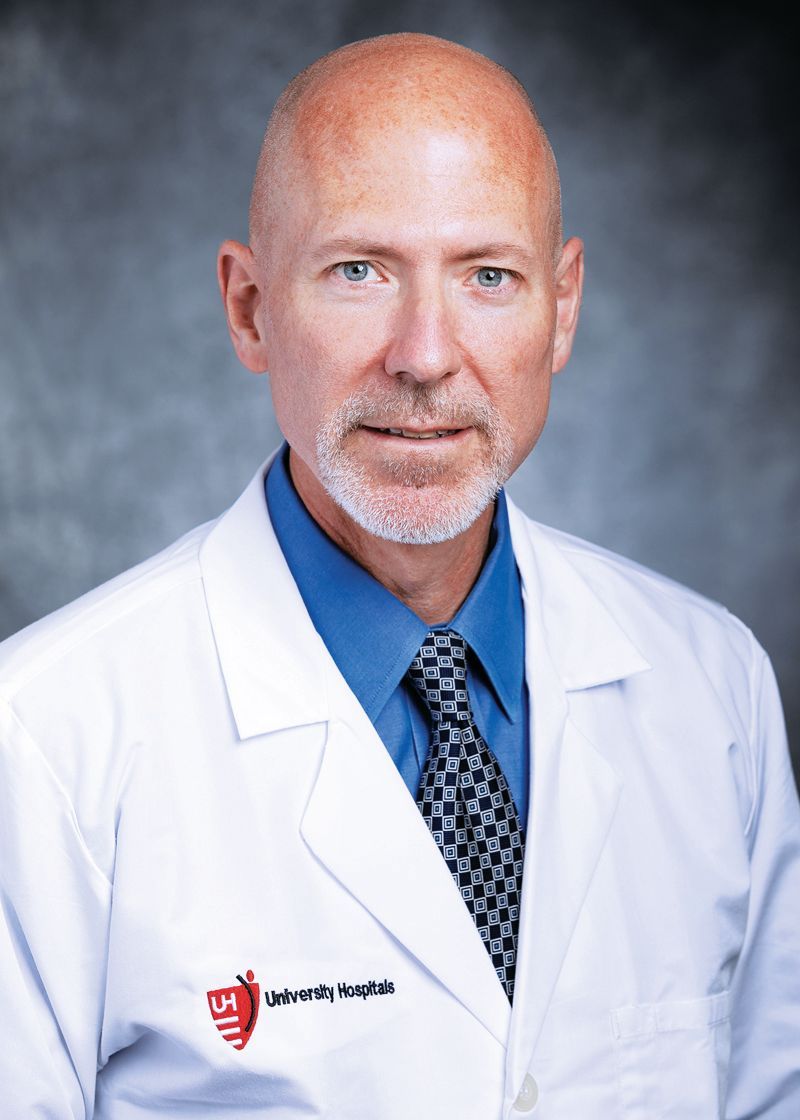 A person in a white lab coat and blue shirt with a patterned tie standing against a gray backdrop.