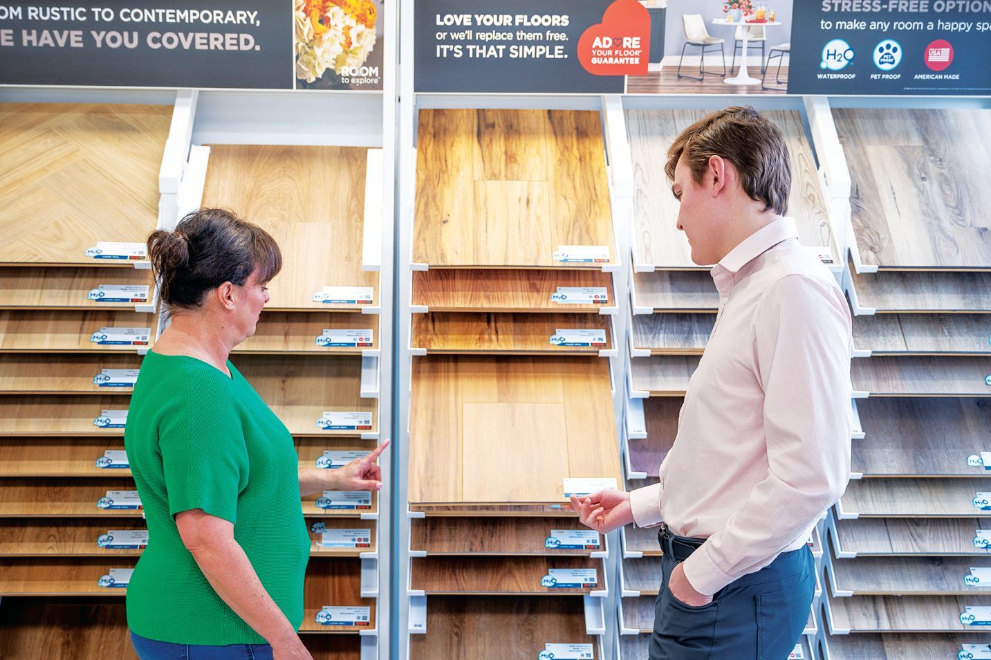 A person in a green shirt and a person in a light shirt examine various wood floor samples in a retail store display.