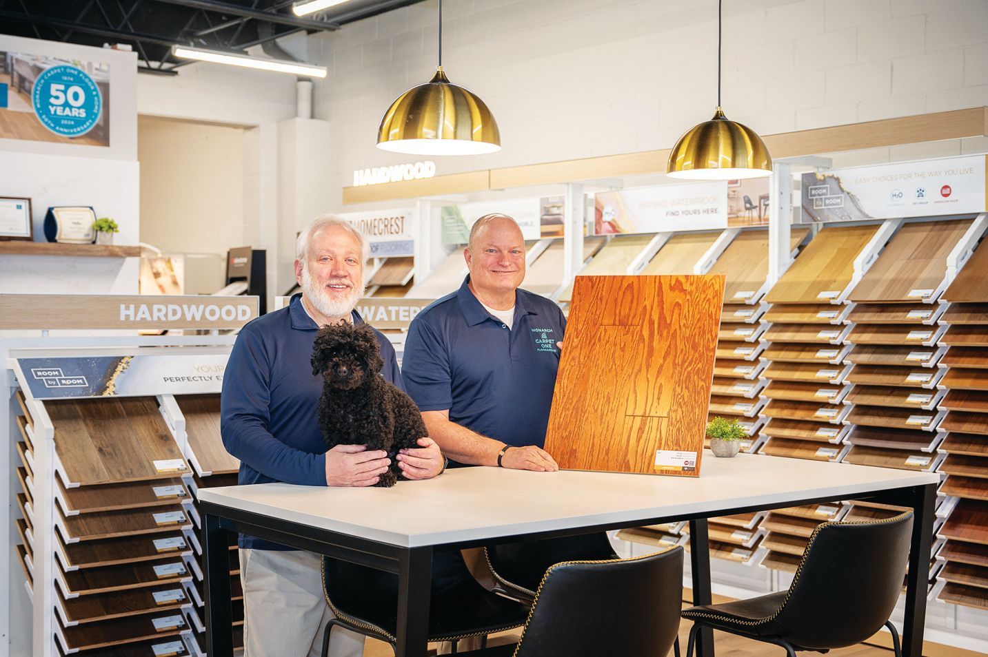 Two professionals and a small dog stand at a table in a flooring showroom, displaying a wooden board sample.