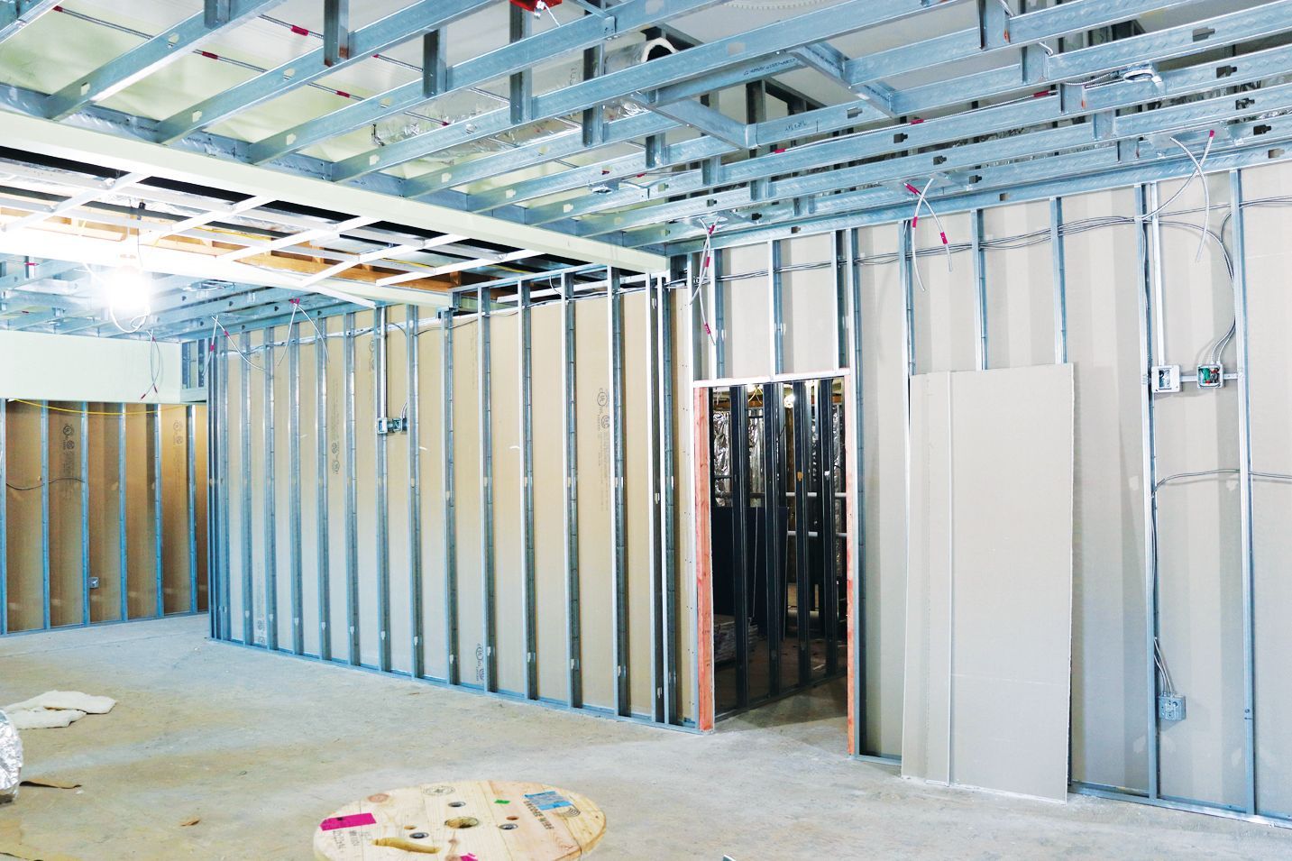 Interior view of a construction site showing metal wall studs, drywall panels, and suspended metal ceiling framing.