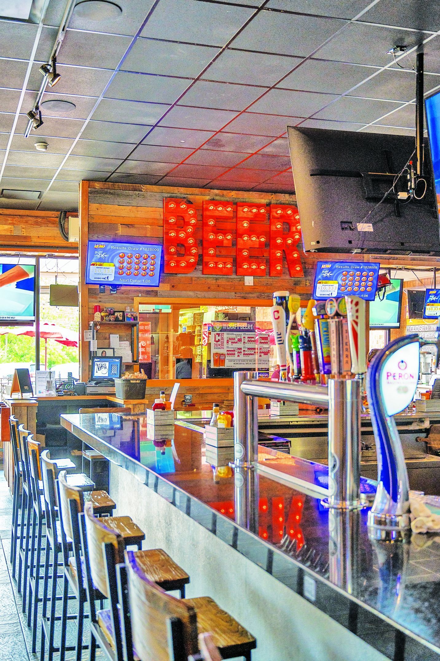 An empty bar interior featuring wooden walls, a long dark countertop, rows of stools, and a glowing neon beer sign.