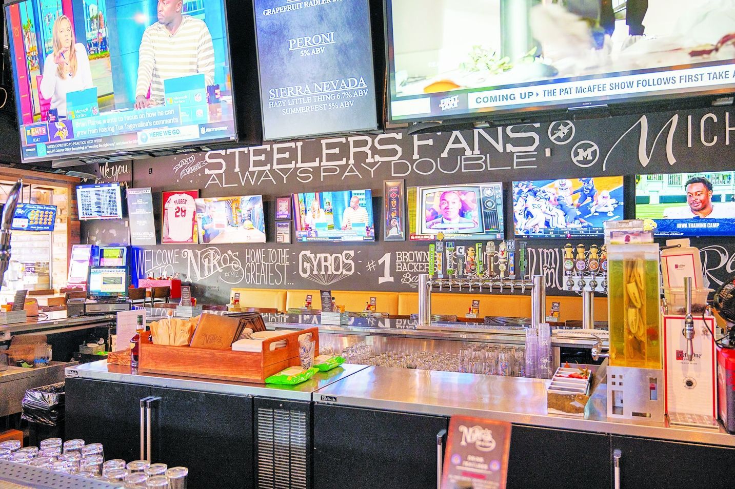 A bar interior with multiple television screens showing sports, a drink station, and a chalkboard sign about Steelers fans.