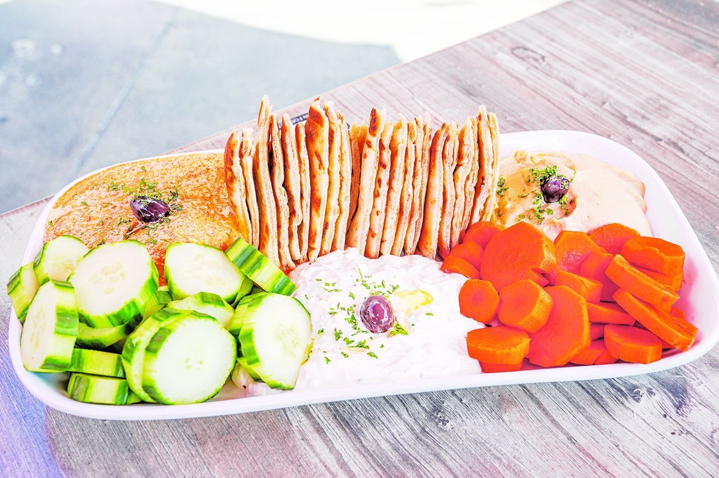 A platter with hummus, tzatziki, pita bread, sliced cucumbers, and sliced carrots on a light wooden table.