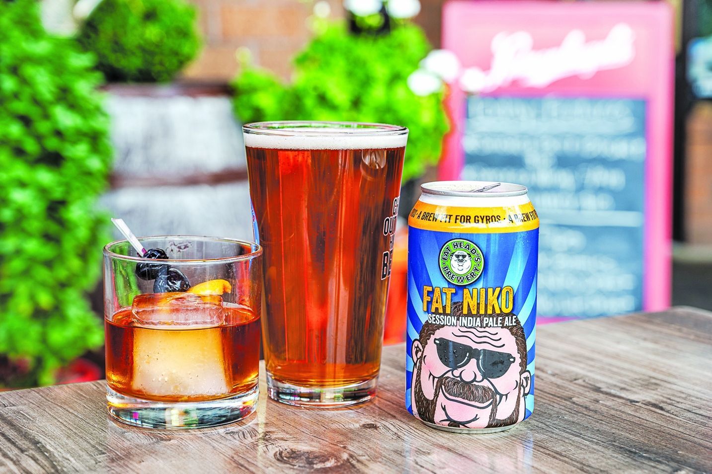 A cocktail, a pint of amber beer, and a Fat Niko beer can sit on an outdoor wooden table with greenery in the background.