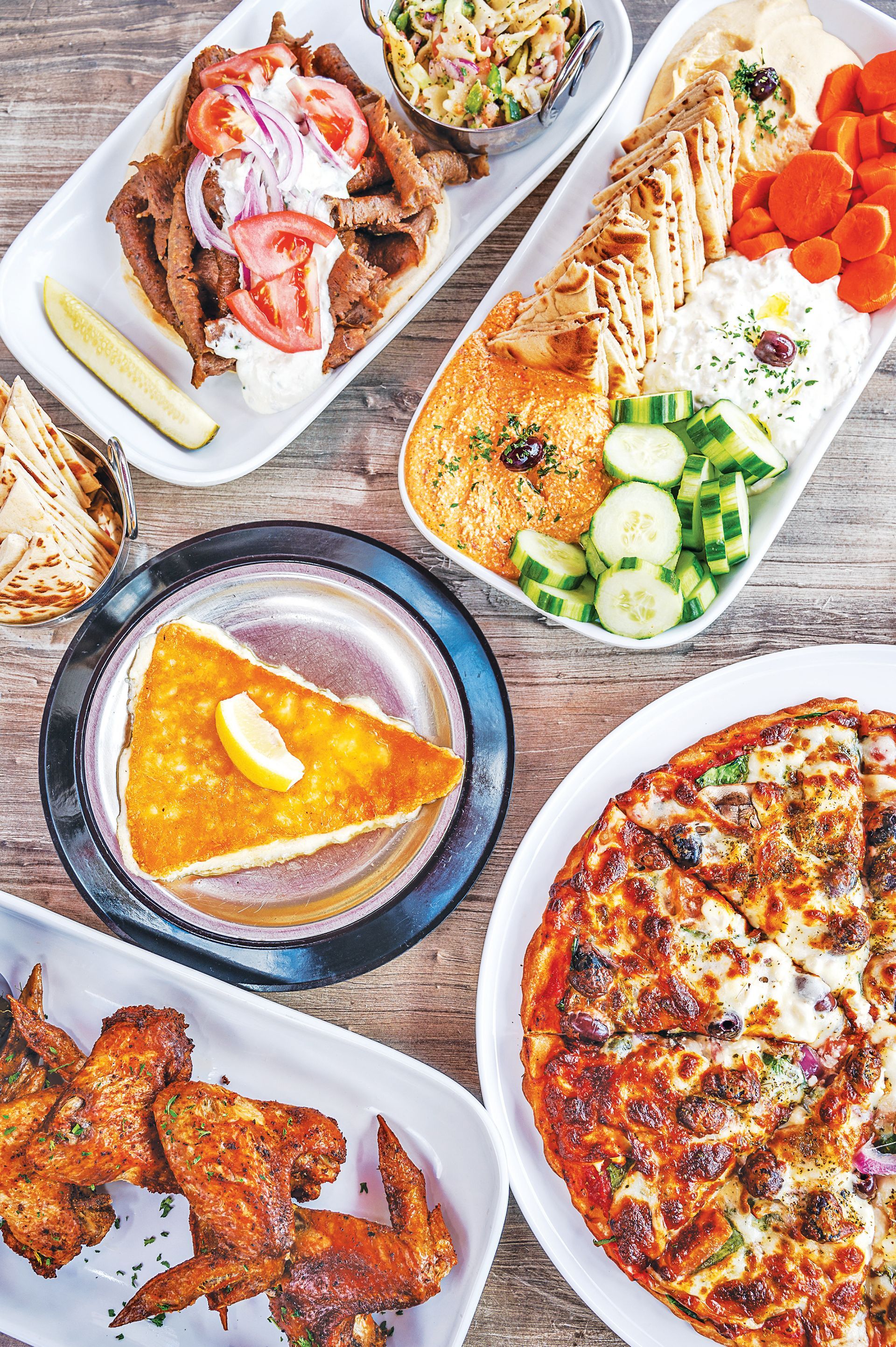 A top-down view of a rustic table with a spread of Mediterranean food, including a pizza, wings, gyros, and appetizers.