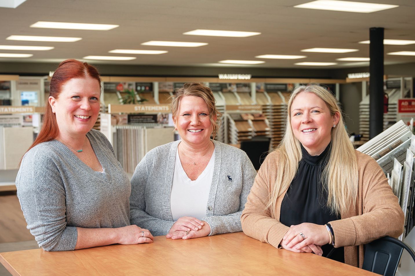Three smiling people stand behind a wooden table in a showroom with flooring samples in the background.
