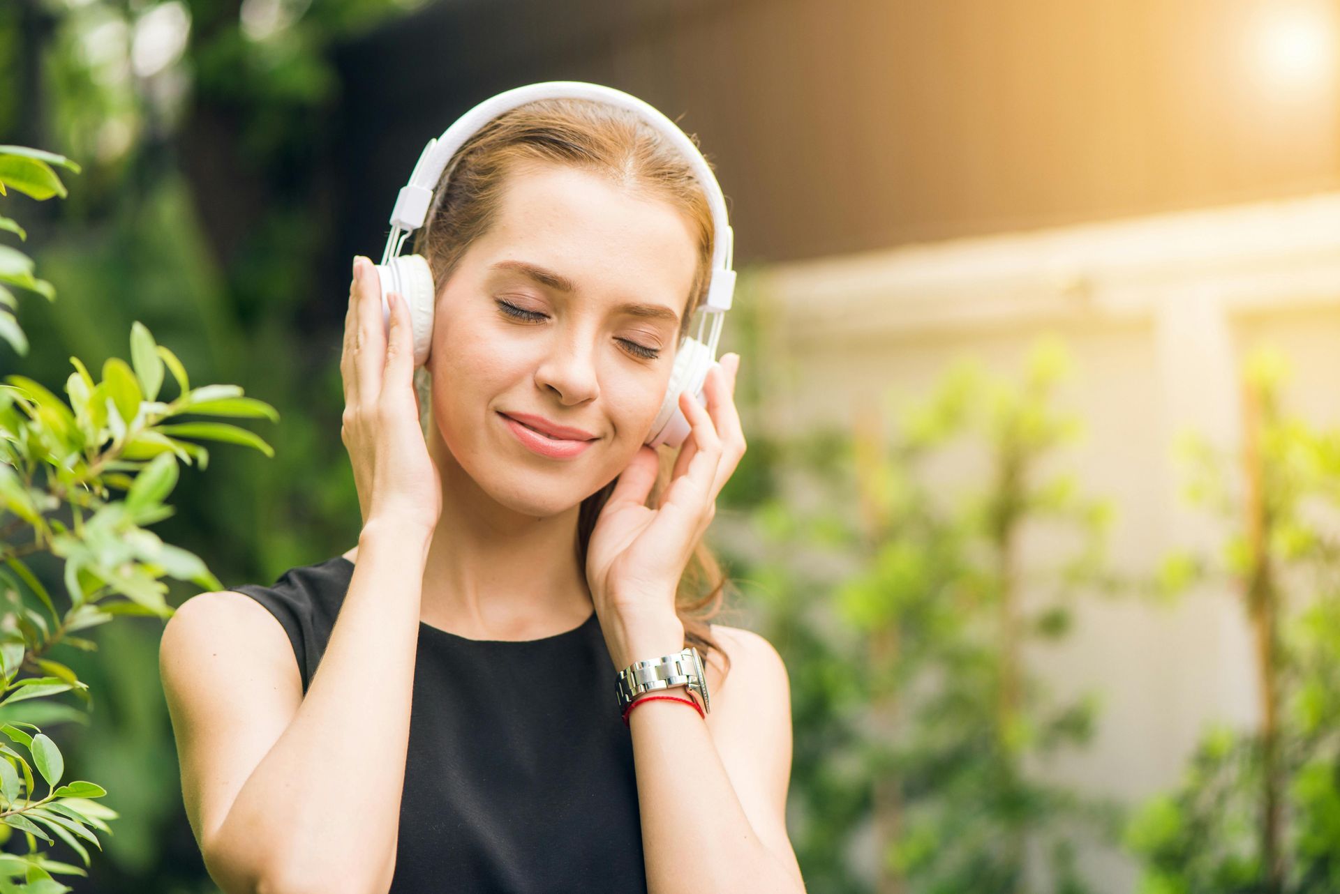 A person with closed eyes wearing white headphones and a black top, smiling while standing outdoors among green plants.