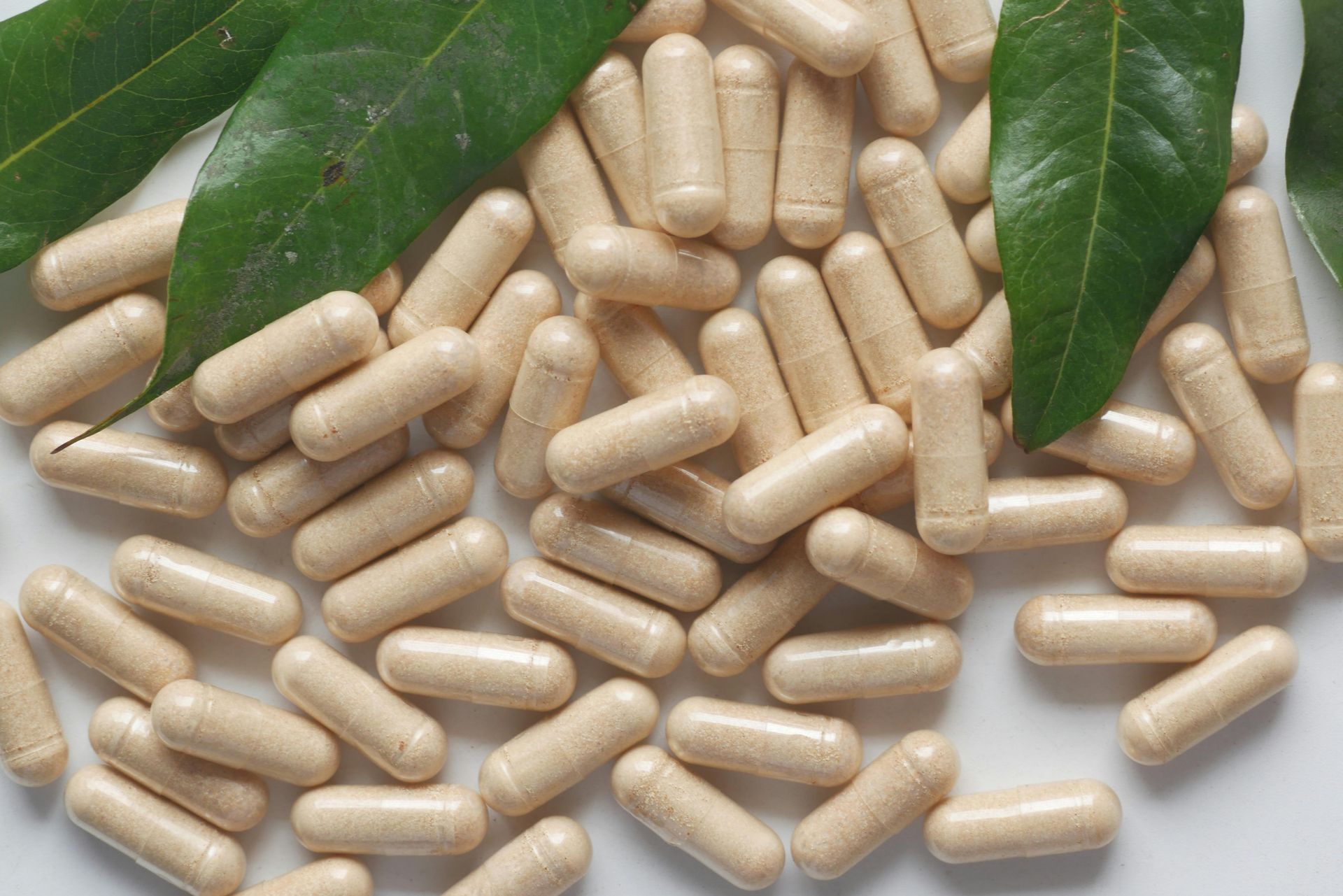 A pile of beige supplement capsules arranged next to green leaves on a white background.