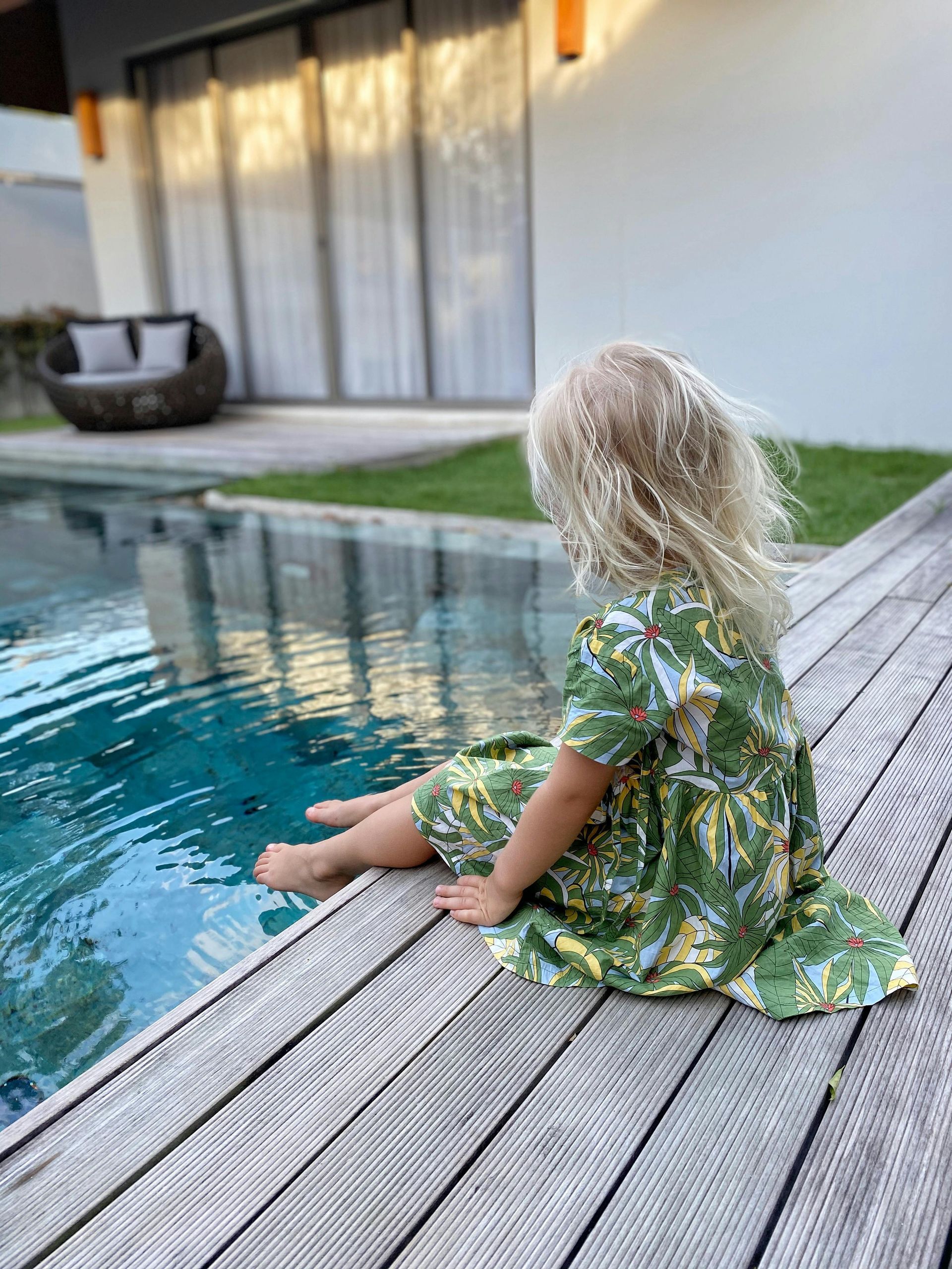 A small child in a green patterned dress sits on a wooden deck by a swimming pool, facing the water.