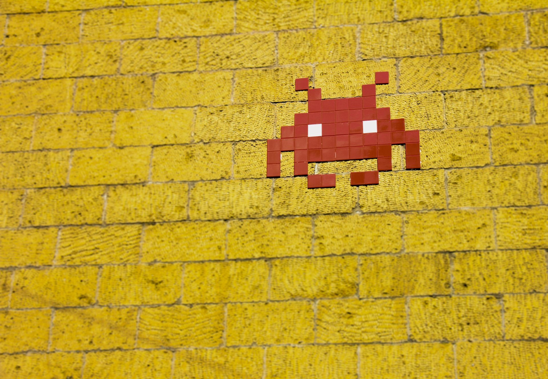 A red mosaic Space Invader tile art piece centered on a bright yellow brick wall.