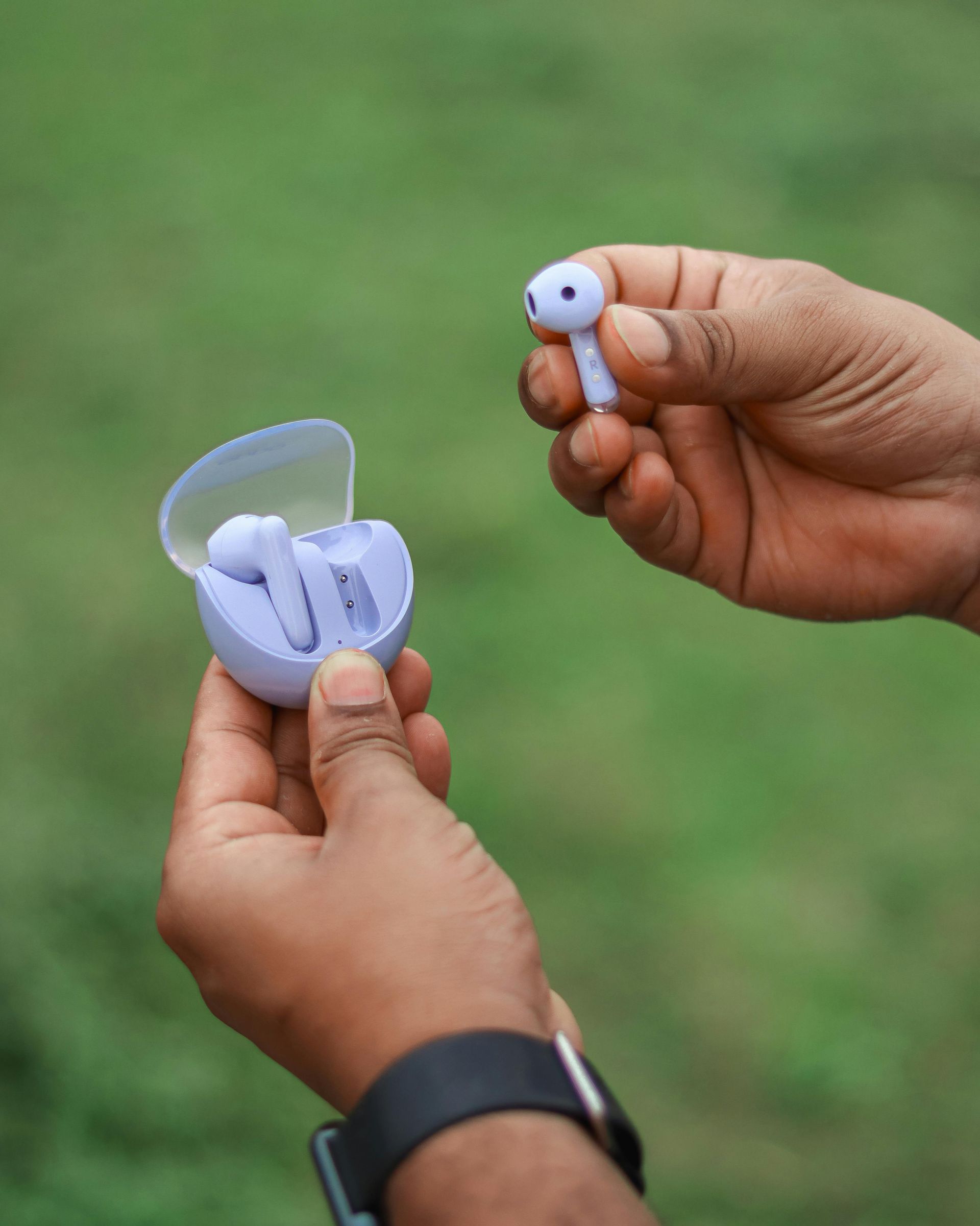 A person holds a lavender-colored wireless earbud case and a single matching earbud outdoors against a blurred green background.