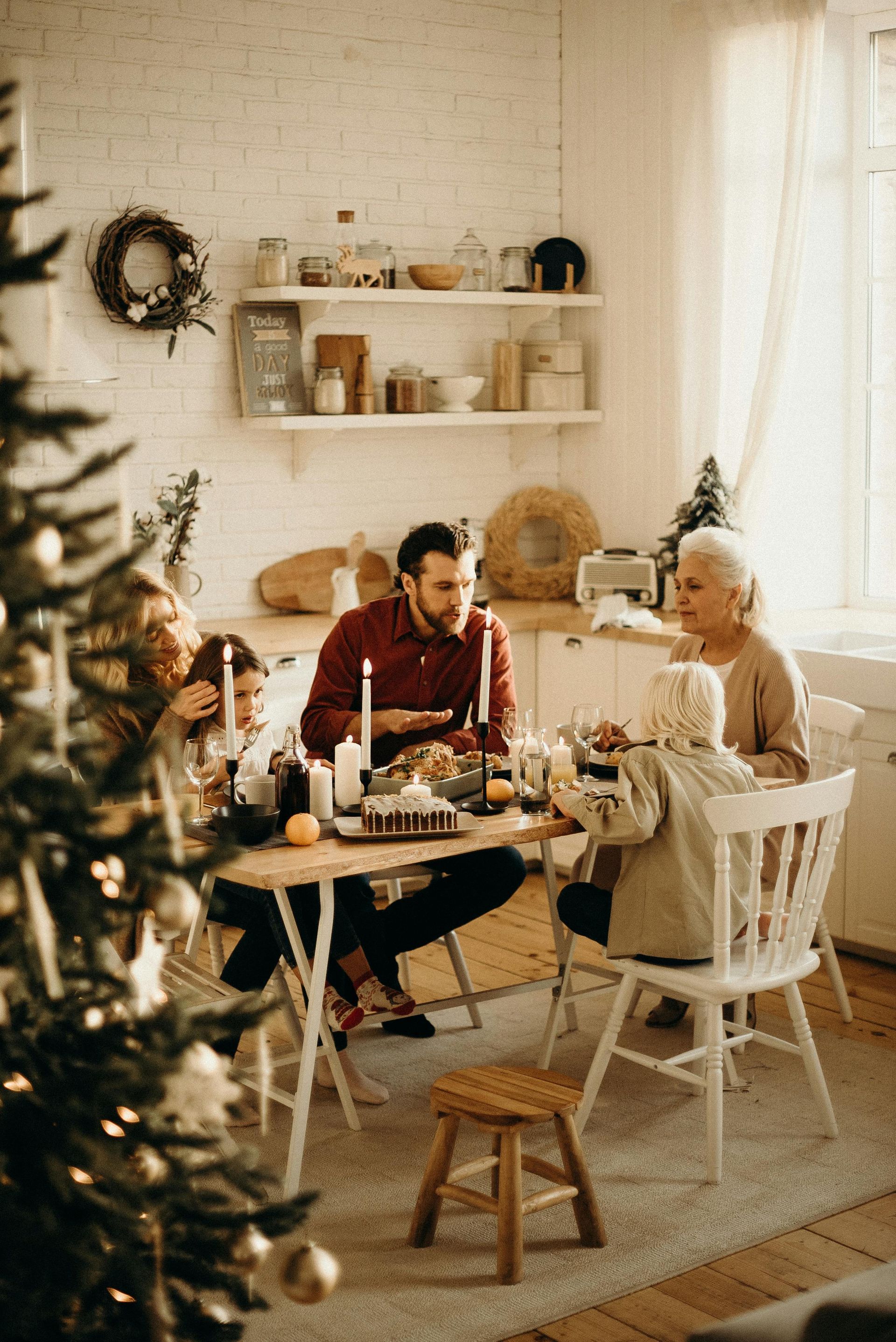 A family sits at a wooden dining table in a cozy, decorated kitchen, enjoying a holiday meal together.