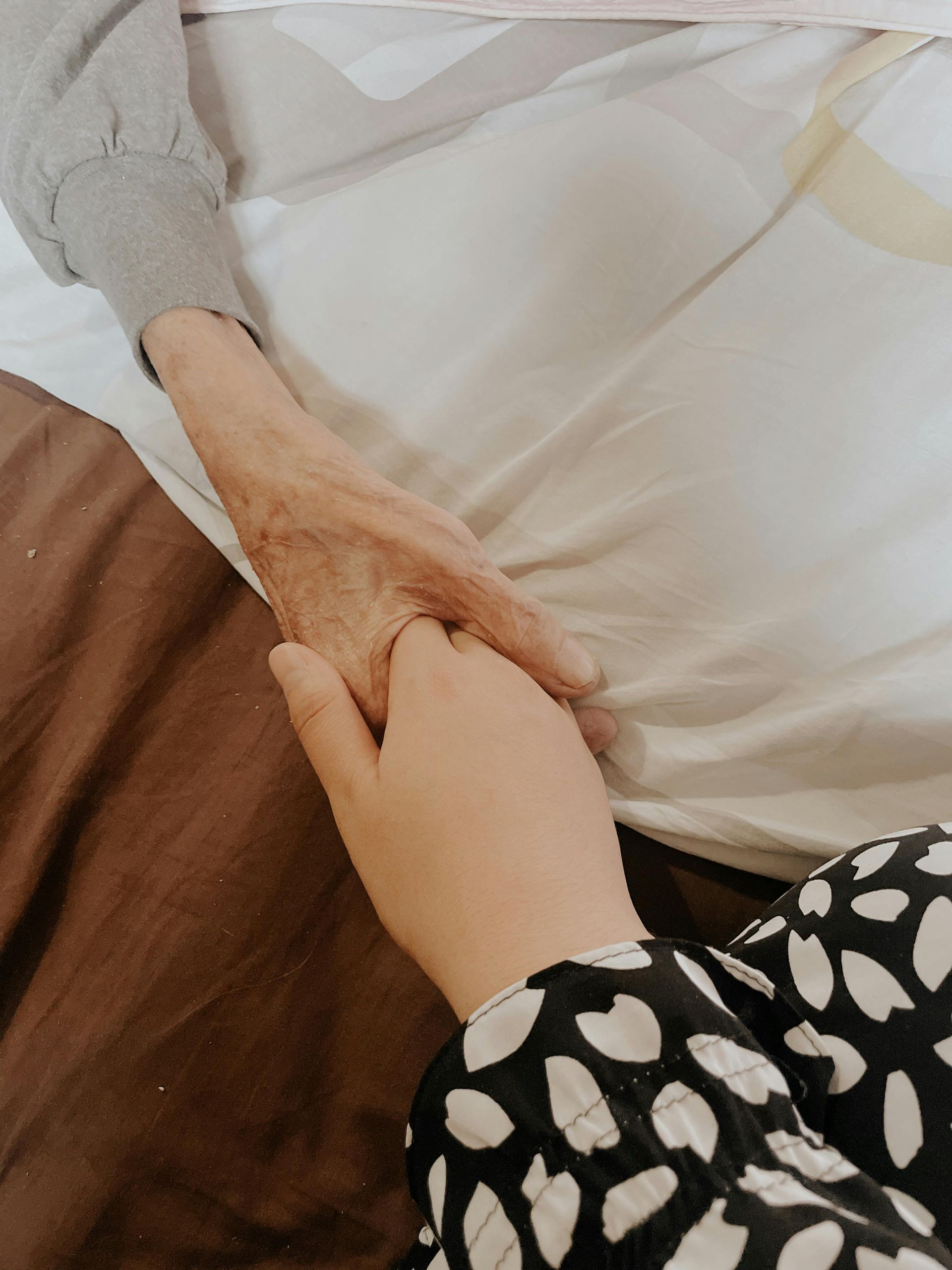 A person’s hand gently holding an older hand in a bed, symbolizing care and connection.